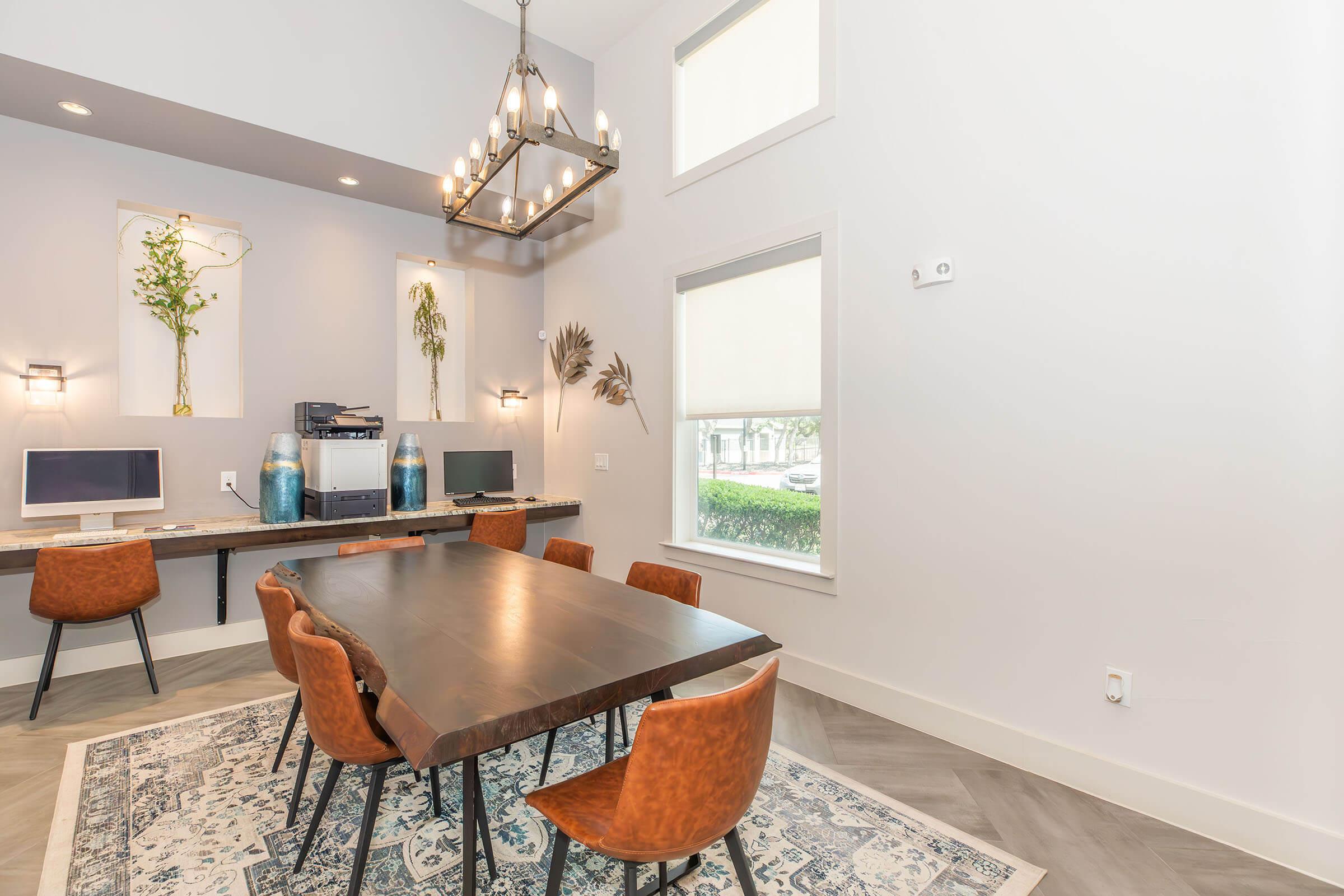 A modern dining area with a wooden table surrounded by six brown leather chairs. The space features a chandelier, two computers on a side table, and decorative plants in wall niches. Large windows allow natural light to brighten the room, complemented by a patterned rug on the floor.