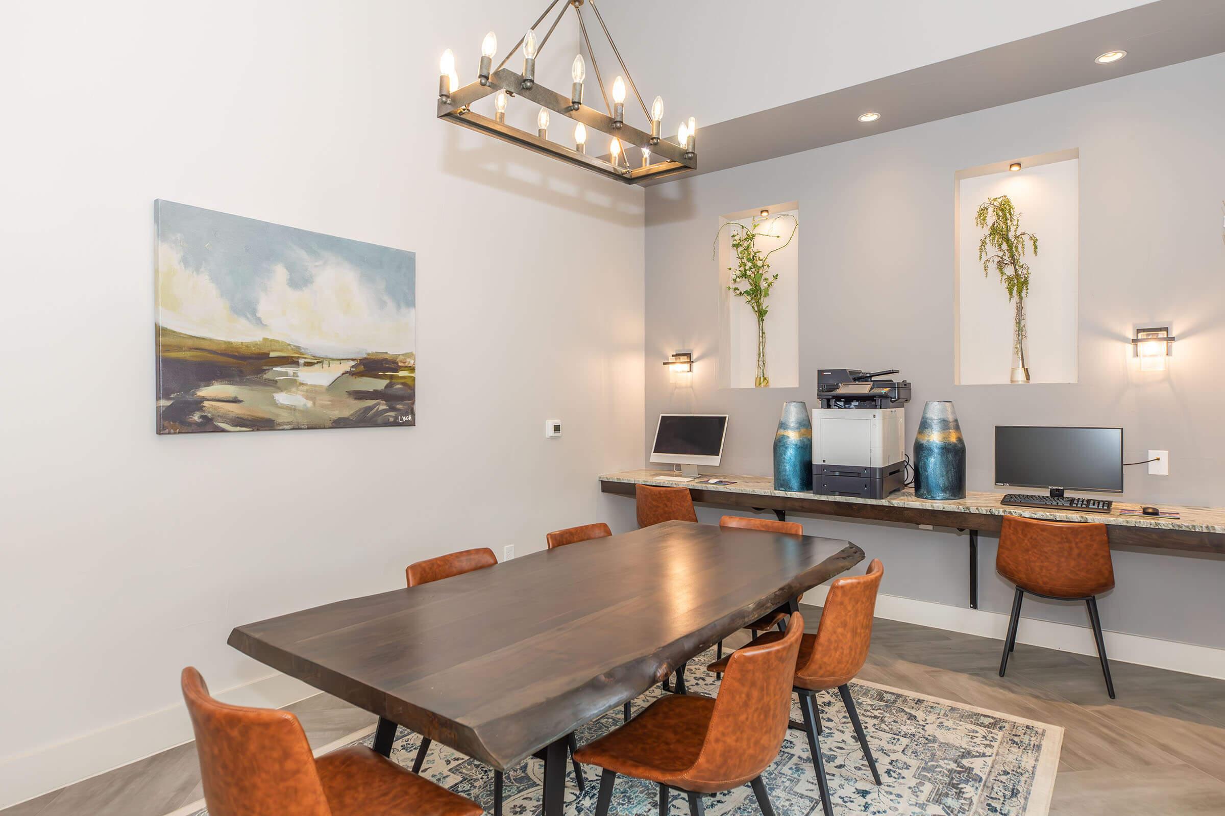 A modern office space featuring a wooden dining table surrounded by six brown chairs. On the wall hangs a landscape painting. Two computers are set up on a shelf against the wall, and a printer is nearby. The room is illuminated by a stylish chandelier and has decorative plants in wall niches.