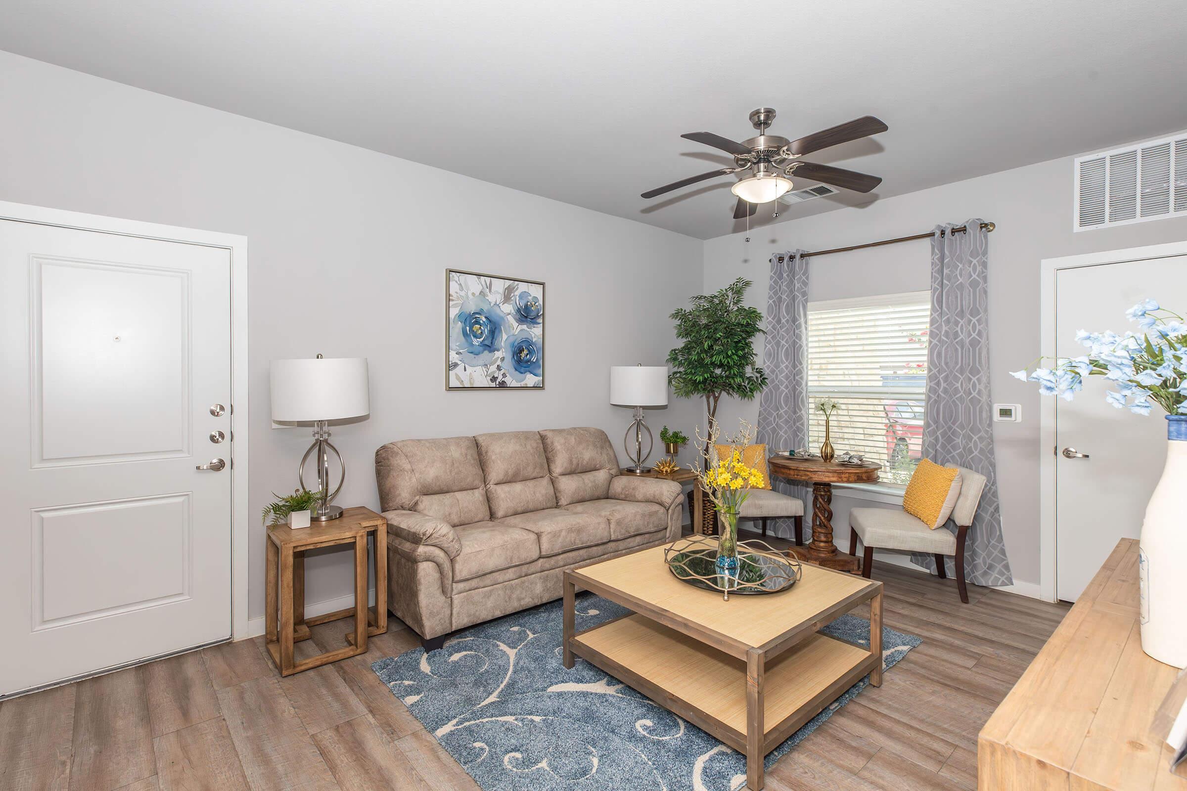 A cozy living room featuring a light-colored sofa, a wooden coffee table, and a decorative blue rug. There are two lamps on side tables, a potted plant, and a small dining table by the window adorned with flowers. The walls are painted in soft tones, complemented by blue wall art.