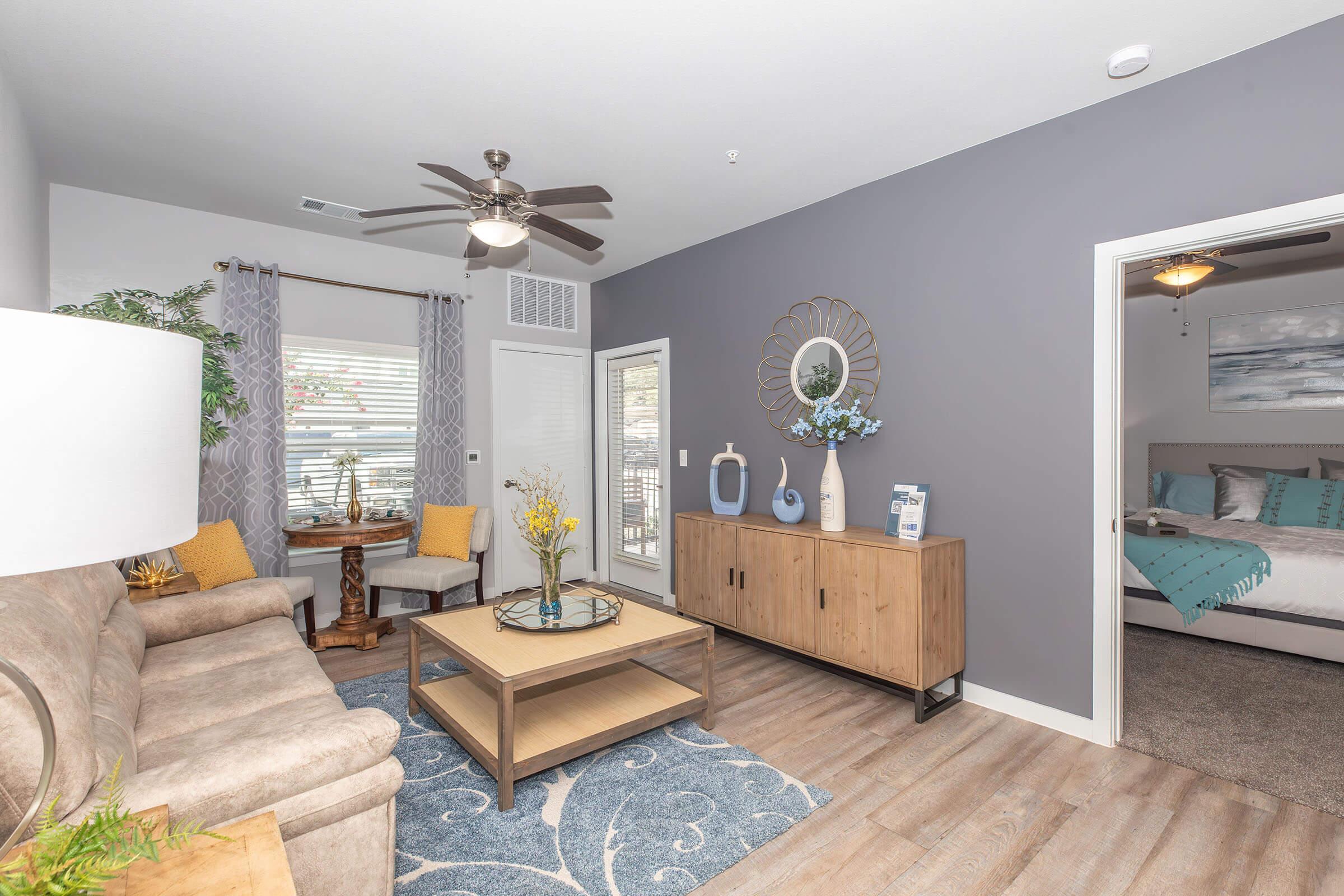 A modern living room featuring a beige sofa, a wooden coffee table, and decorative items on a sideboard. There are yellow accent pillows, potted plants, and large windows allowing natural light. A doorway leads to a bedroom area, and the walls are painted in a soft gray tone.