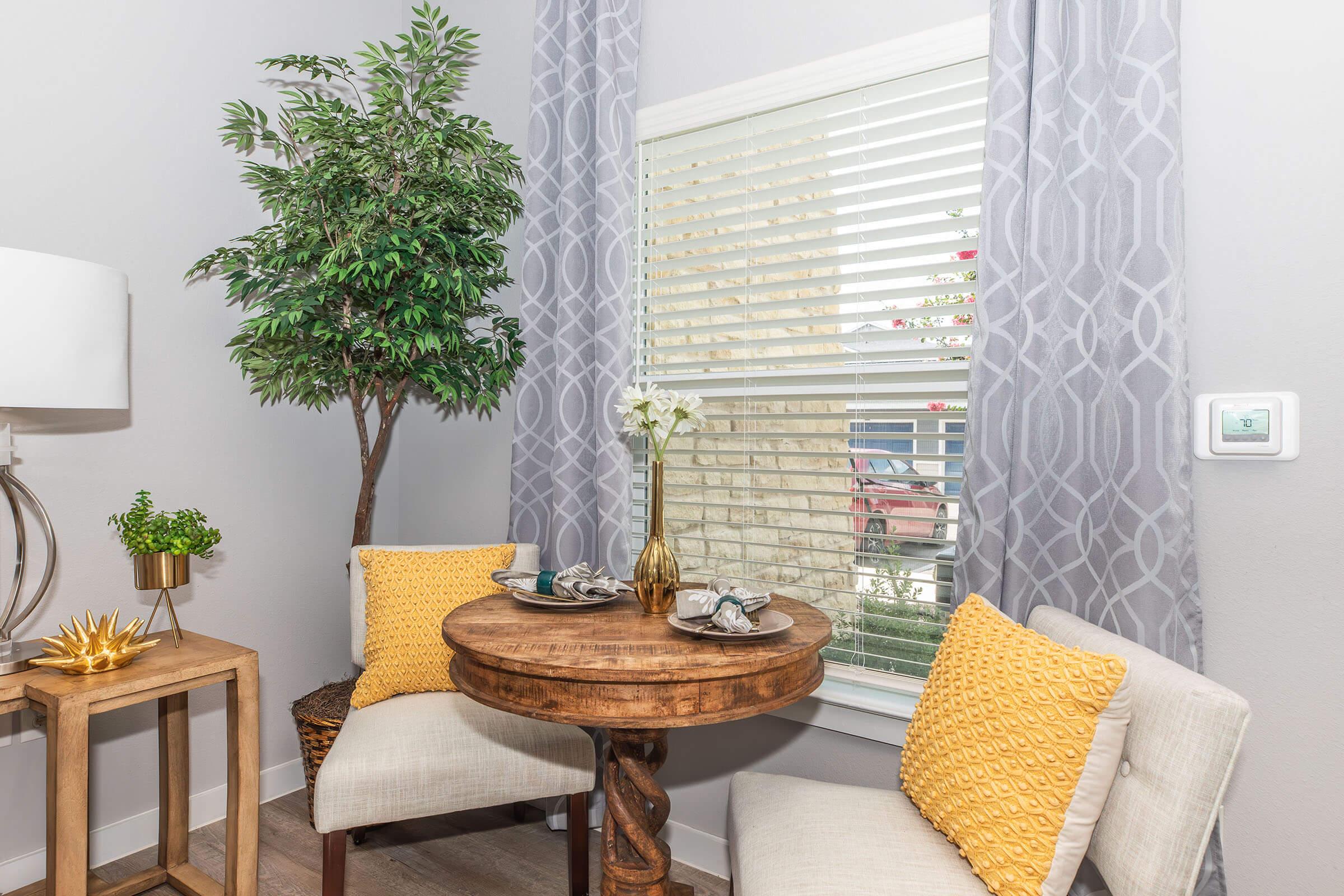 A cozy dining nook featuring a round wooden table set for two, with decorative plates and a vase of flowers. Surrounding the area are two cushioned chairs with yellow pillows, a small potted tree, and light gray curtains. Natural light streams in through the blinds on a window, creating a warm atmosphere.
