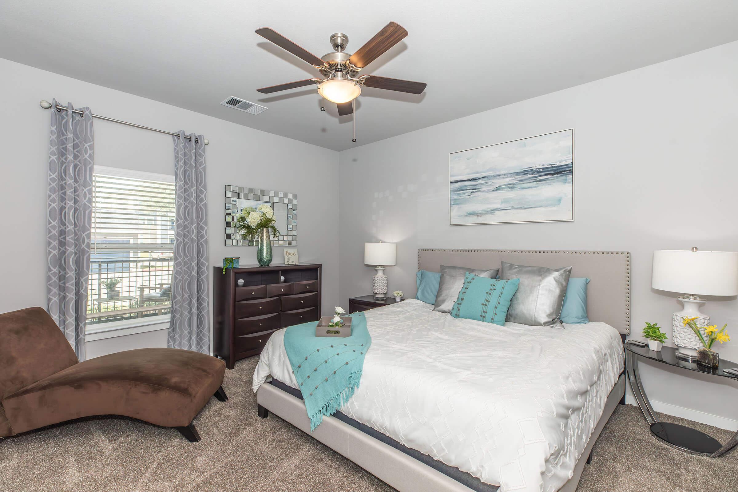 A cozy bedroom featuring a queen-sized bed with light gray bedding and decorative teal pillows. There's a dark wooden dresser, a stylish mirror, and a small side table with lamps. A brown chaise lounge is positioned to one side, and large windows with gray curtains allow natural light to brighten the space.