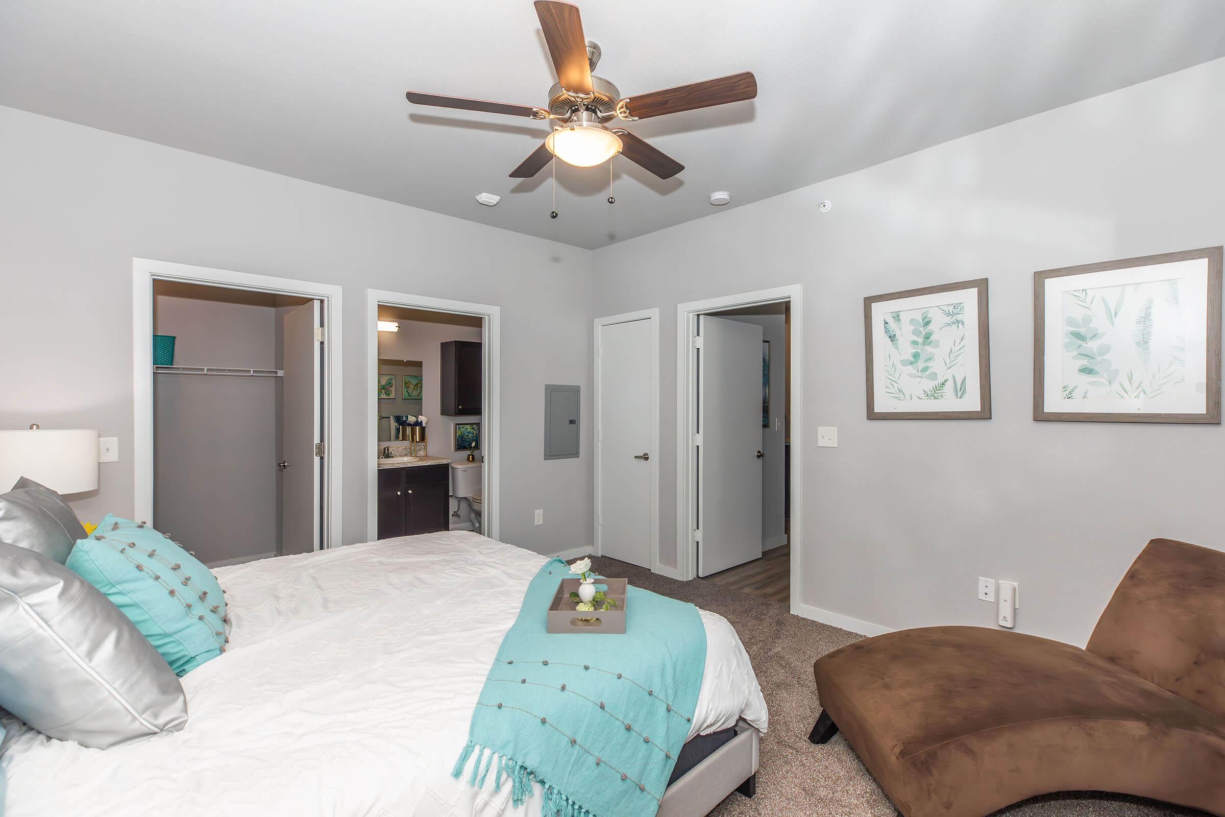 A modern bedroom featuring a queen-sized bed with gray and teal accents, a ceiling fan, and two doors leading to adjacent rooms. A cozy brown chaise lounge is positioned beside the bed, and artwork of green plants adorns the walls. The room has neutral-colored walls and carpet, creating a warm atmosphere.