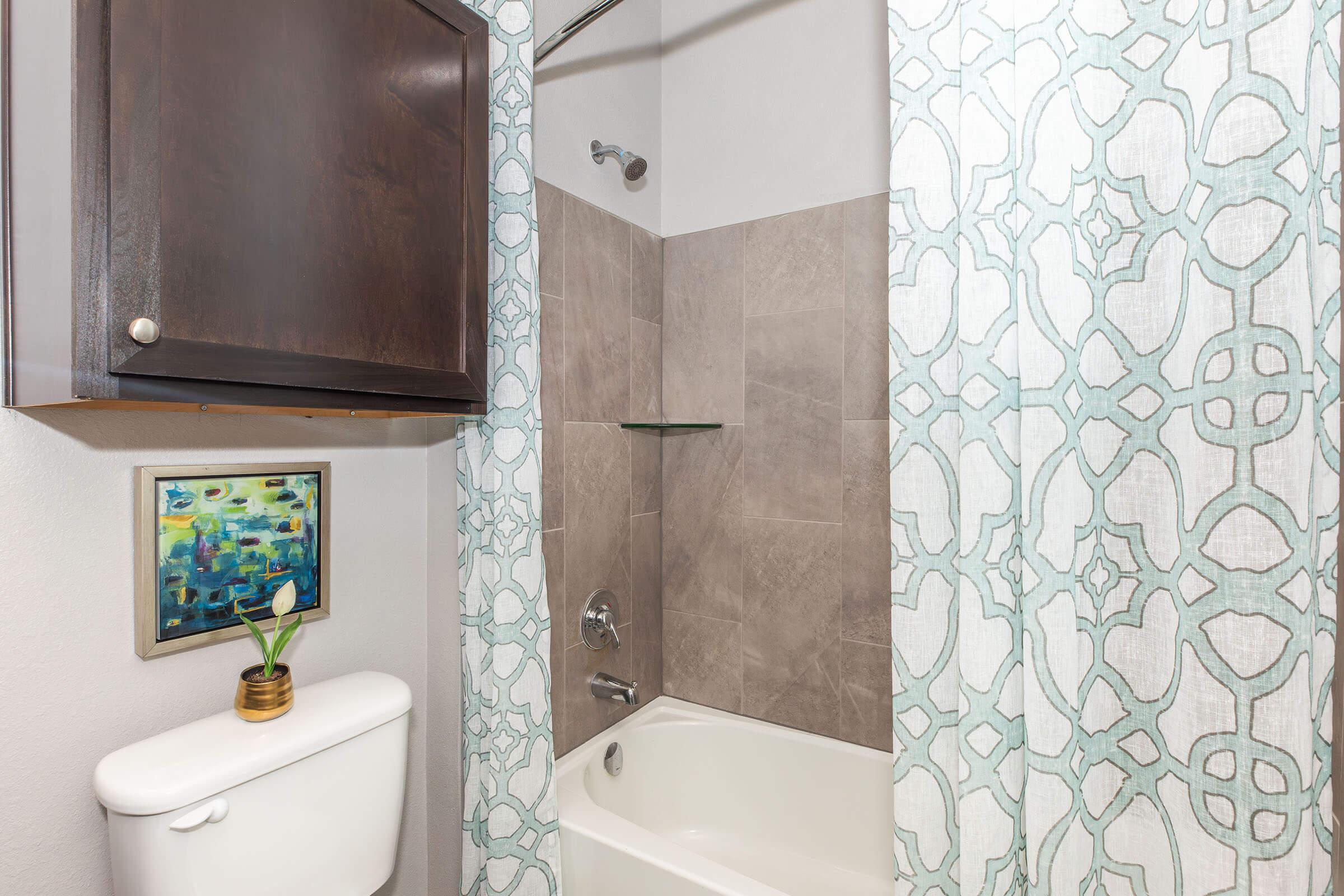 A small bathroom featuring a bathtub with a showerhead. The walls are tiled in a light brown color, and a decorative shower curtain with a teal and white pattern hangs from a rod. A wall-mounted cabinet is above the toilet, and there's a framed piece of artwork beside a small plant on the countertop.