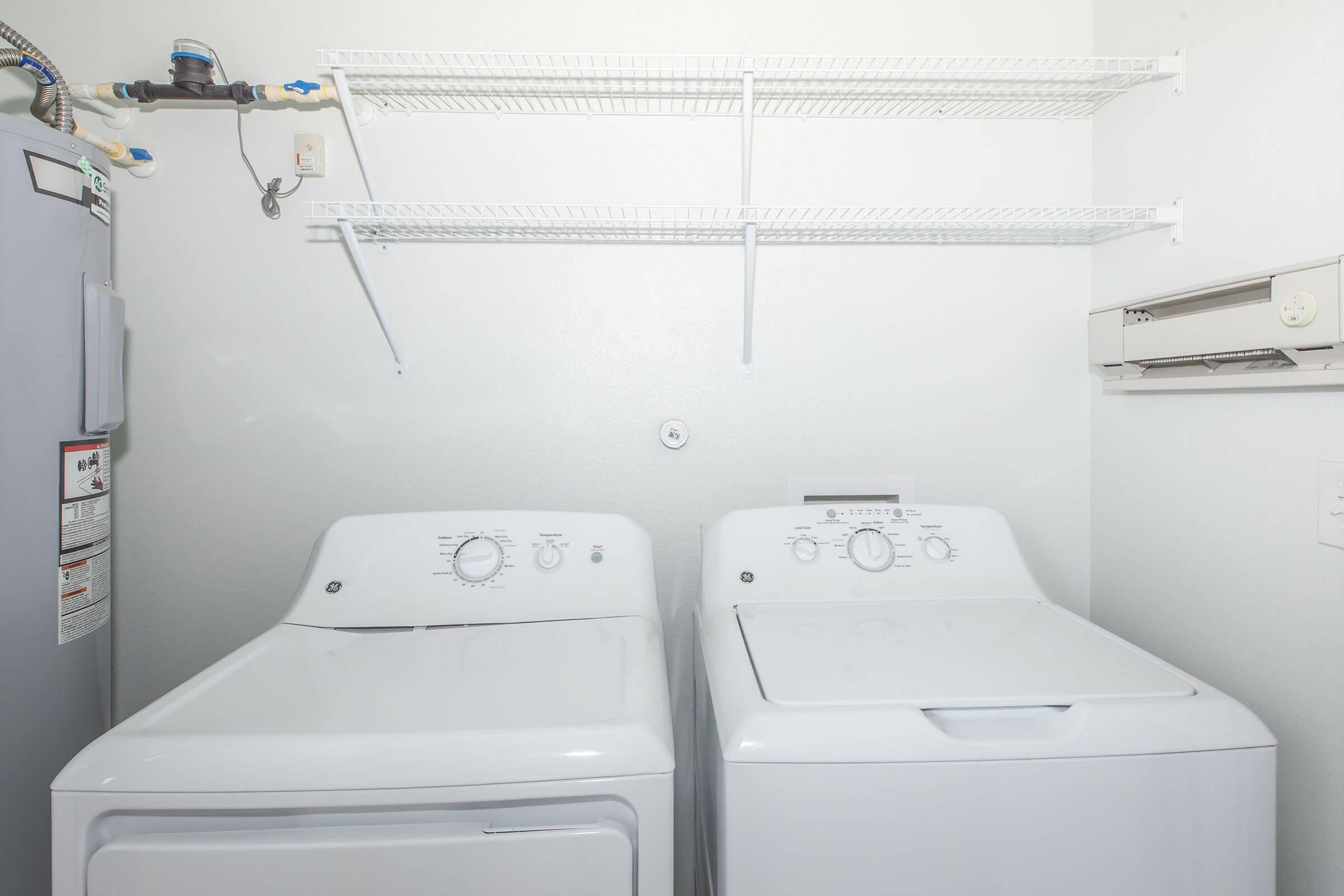 A clean laundry room featuring a white washer and dryer side by side. Above them are wire shelves for storage. The wall is light-colored, and there is a utility hookup visible next to the appliances, creating a functional and organized laundry space.