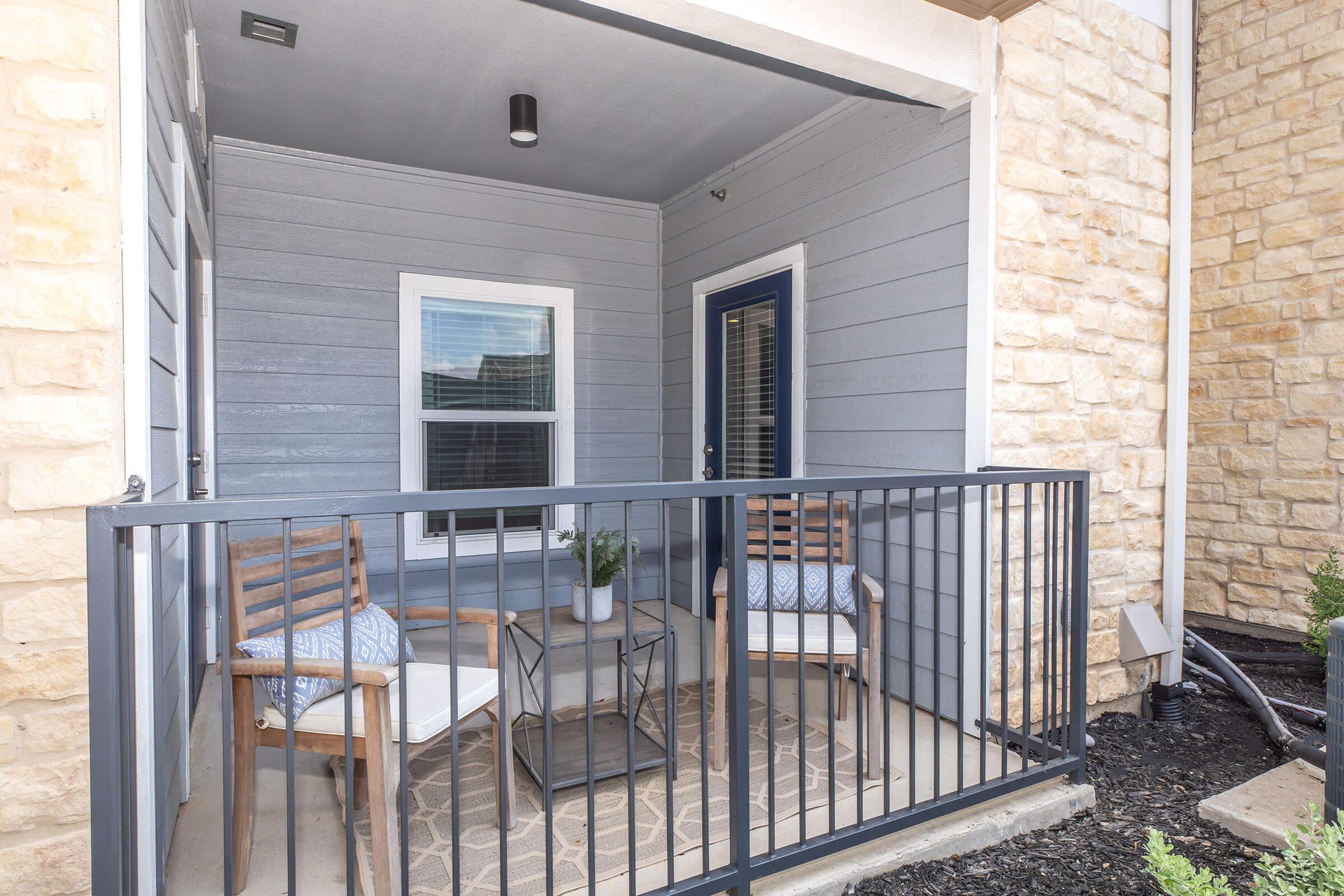 A small balcony or patio area featuring two wooden chairs with blue cushions, a glass table, and a decorative plant. The walls are a mix of stone and blue siding, with a railing enclosing the space. Natural light enters through a window, creating a cozy atmosphere.