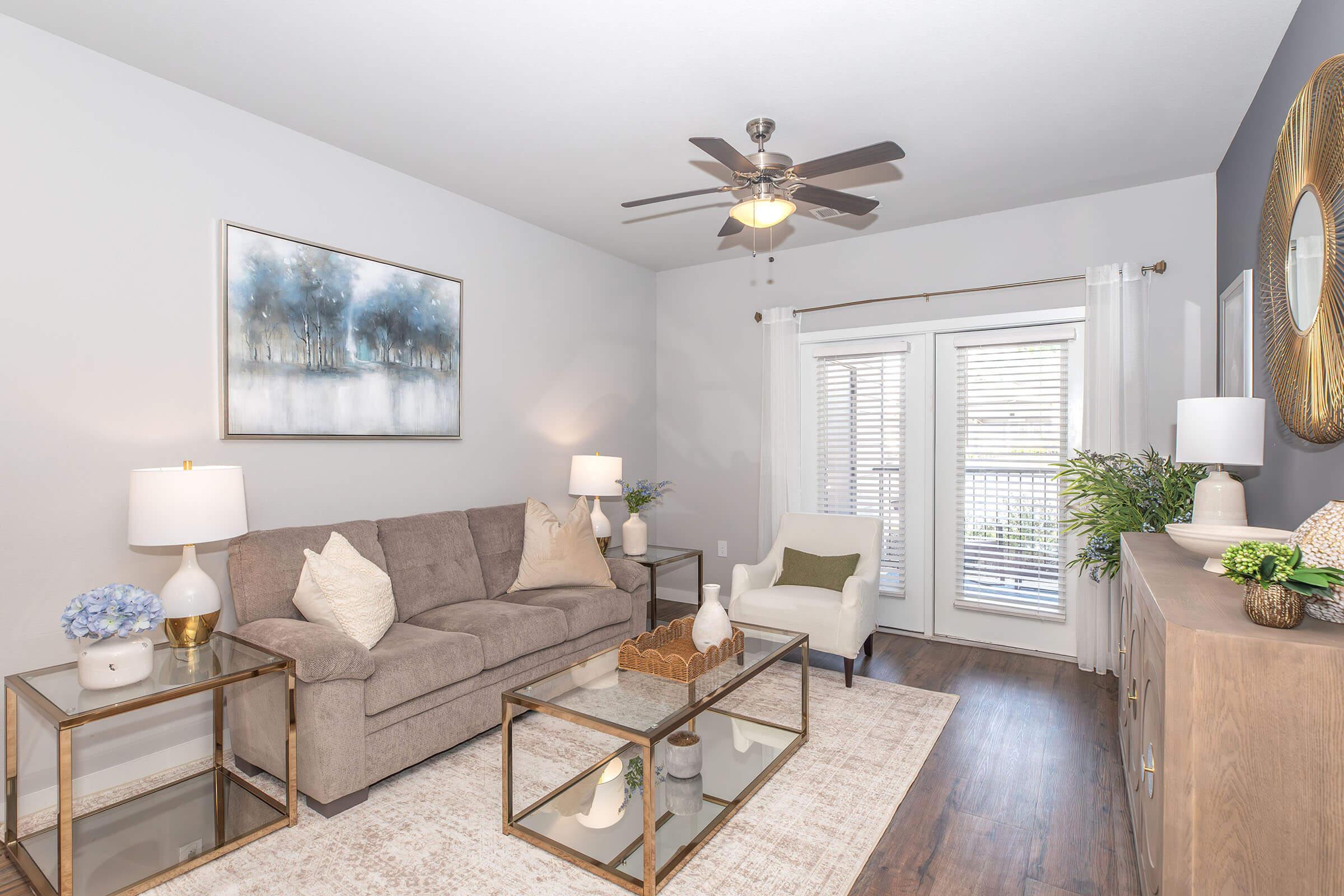 A cozy living room featuring a light gray sofa with decorative pillows, an armchair, and a coffee table with a decorative centerpiece. Two lamps on either side of the sofa provide warm lighting. A large painting of a serene landscape hangs above the couch, and glass doors lead to a small balcony with white shutters.