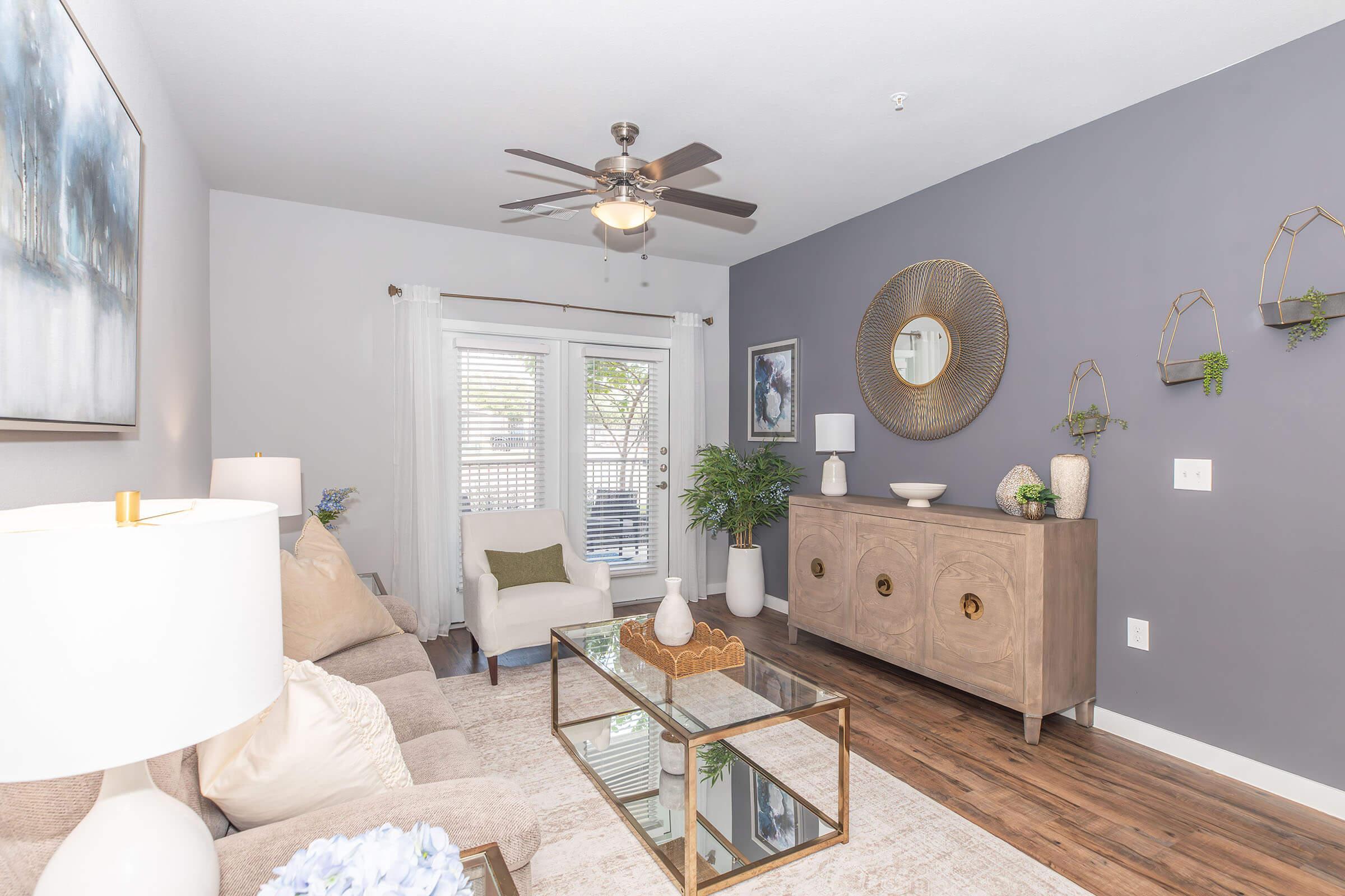 A modern living room featuring a light-colored sofa with decorative cushions, a glass coffee table, and wooden flooring. There is a decorative chest against the wall, potted plants, and wall-mounted planters. A ceiling fan and artwork add a stylish touch, with large windows allowing natural light in.