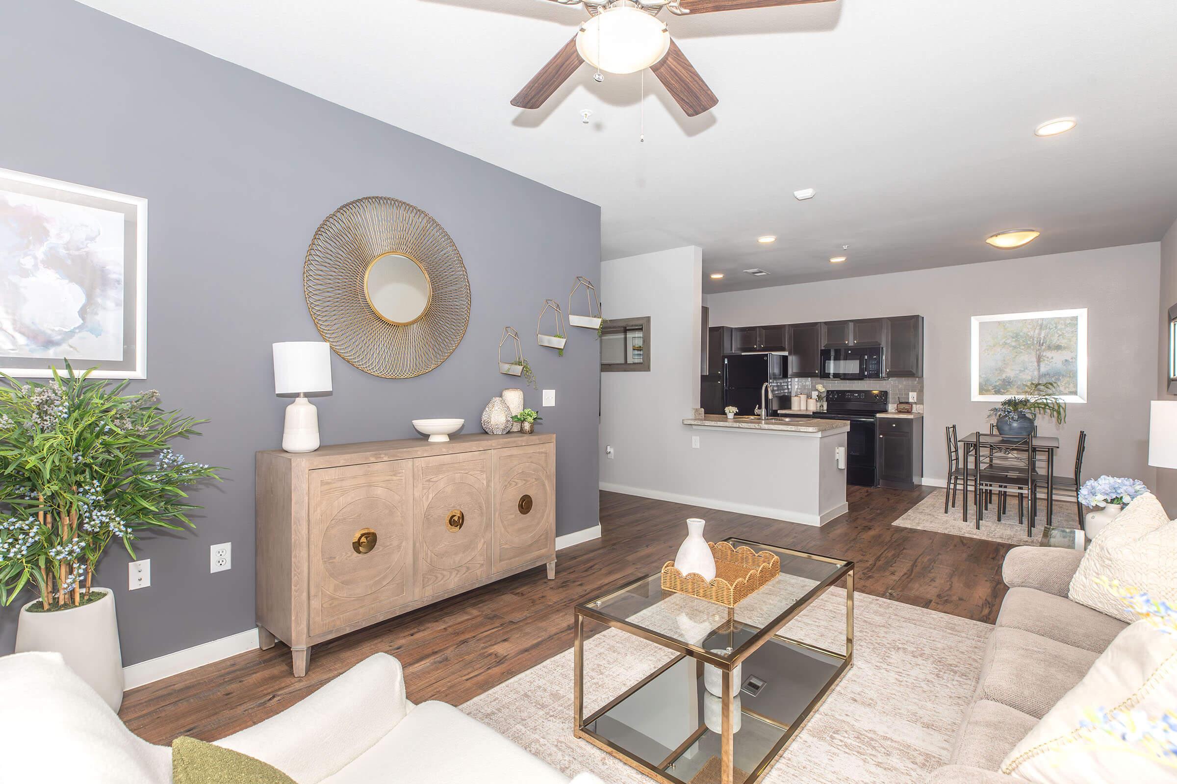 A modern living room featuring a light-colored sofa, a decorative coffee table, and a sideboard with decor items. The walls are painted light gray, and there is a ceiling fan above. In the background, an open kitchen with dark cabinetry is visible, along with a small dining area. Green plants add a touch of nature to the space.