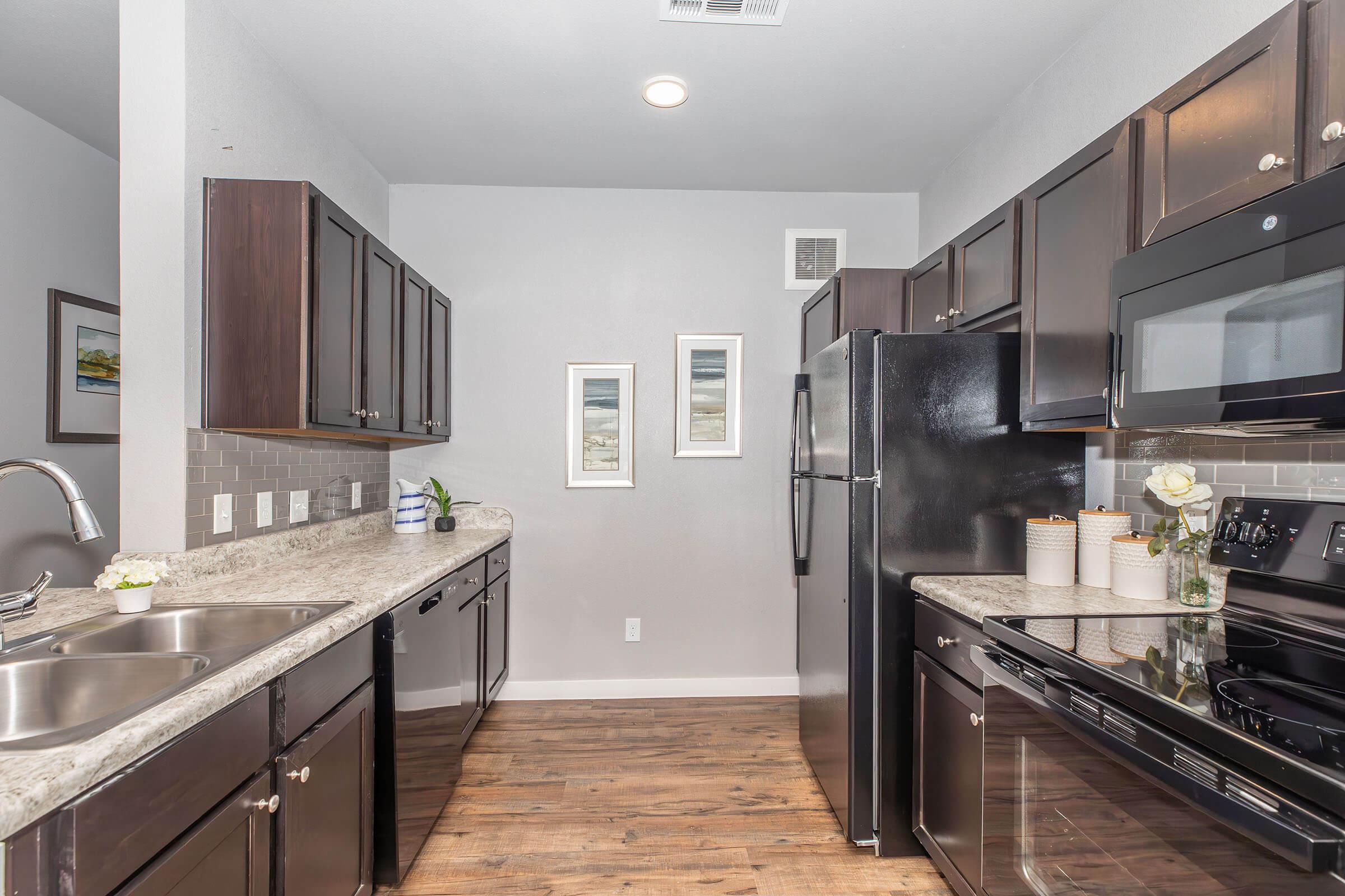 Modern kitchen featuring dark wooden cabinets, stainless steel appliances, and a granite countertop. The layout includes a double sink, electric stove, and a large refrigerator, with framed artwork on the walls and a small decorative plant on the counter. The flooring is wood-like laminate.