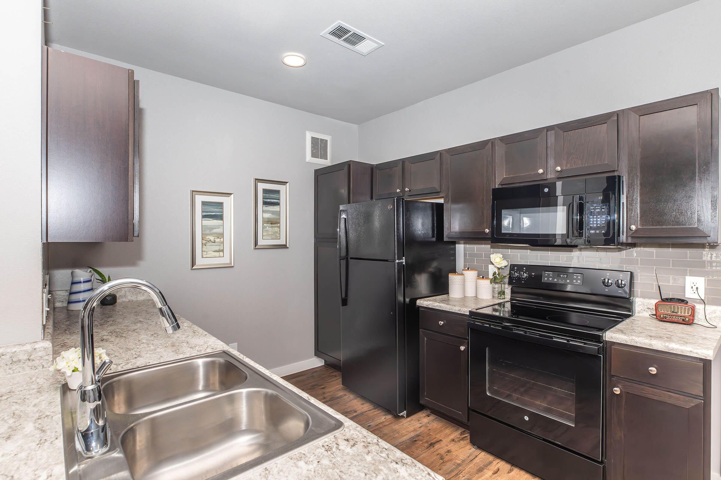 Modern kitchen featuring dark wood cabinets, a black refrigerator, microwave, and stove. The countertop is made of light-colored stone, and there are two framed pictures on the wall. The floor has a warm wood finish, and there are small decorative items on the countertop. Bright, well-lit space.