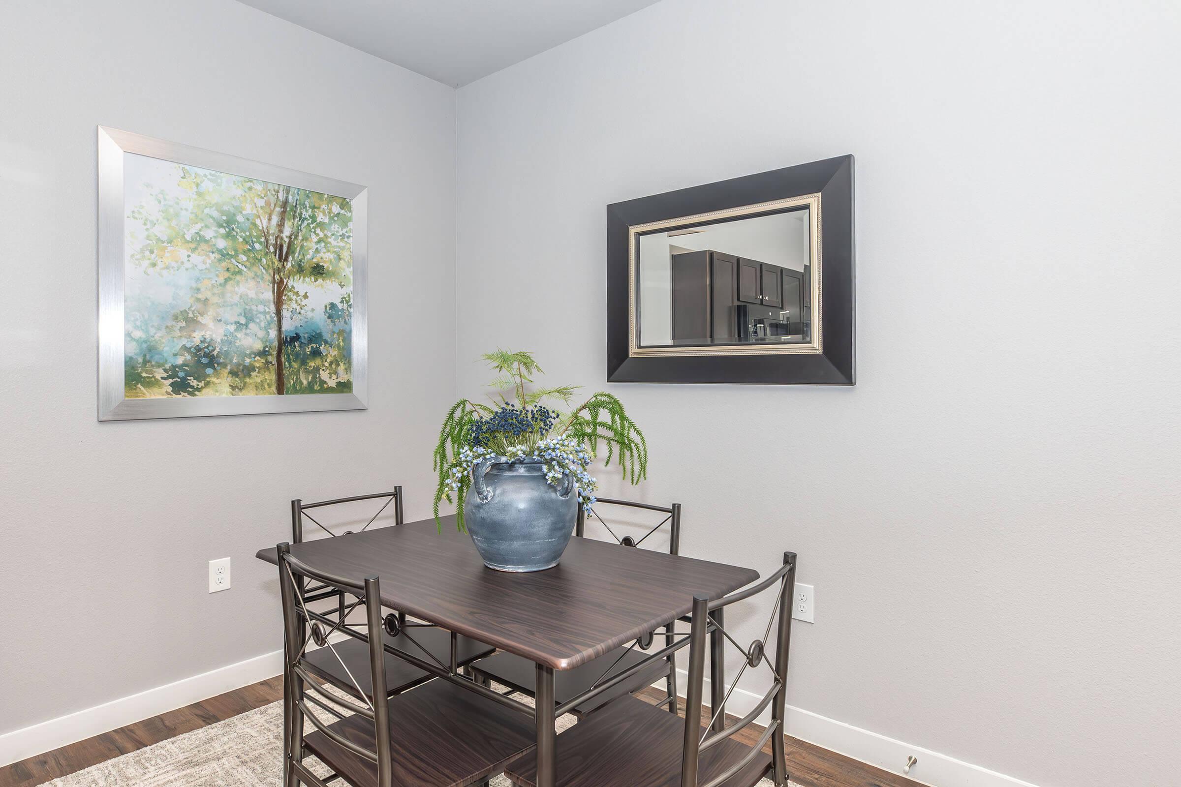 A modern dining area featuring a dark wooden table with four black chairs. A large blue vase with greenery sits on the table. The walls are painted light gray, adorned with a colorful landscape painting and a framed mirror. Hardwood flooring completes the contemporary look.