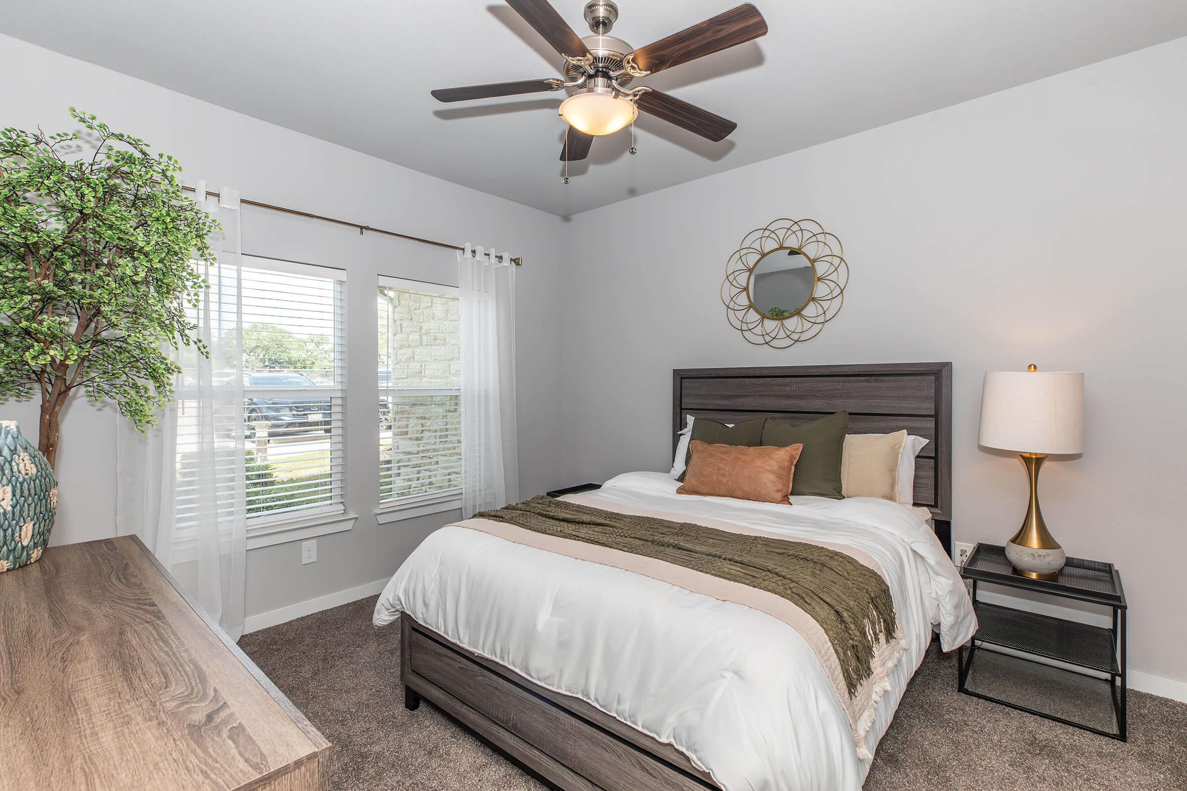 A cozy bedroom featuring a bed with white and olive green linens, a decorative throw pillow, and a wooden headboard. The room has a ceiling fan and a lamp on a nightstand. Natural light streams in through two windows with sheer curtains, complemented by a potted plant and a wall mirror. The decor is modern and inviting.
