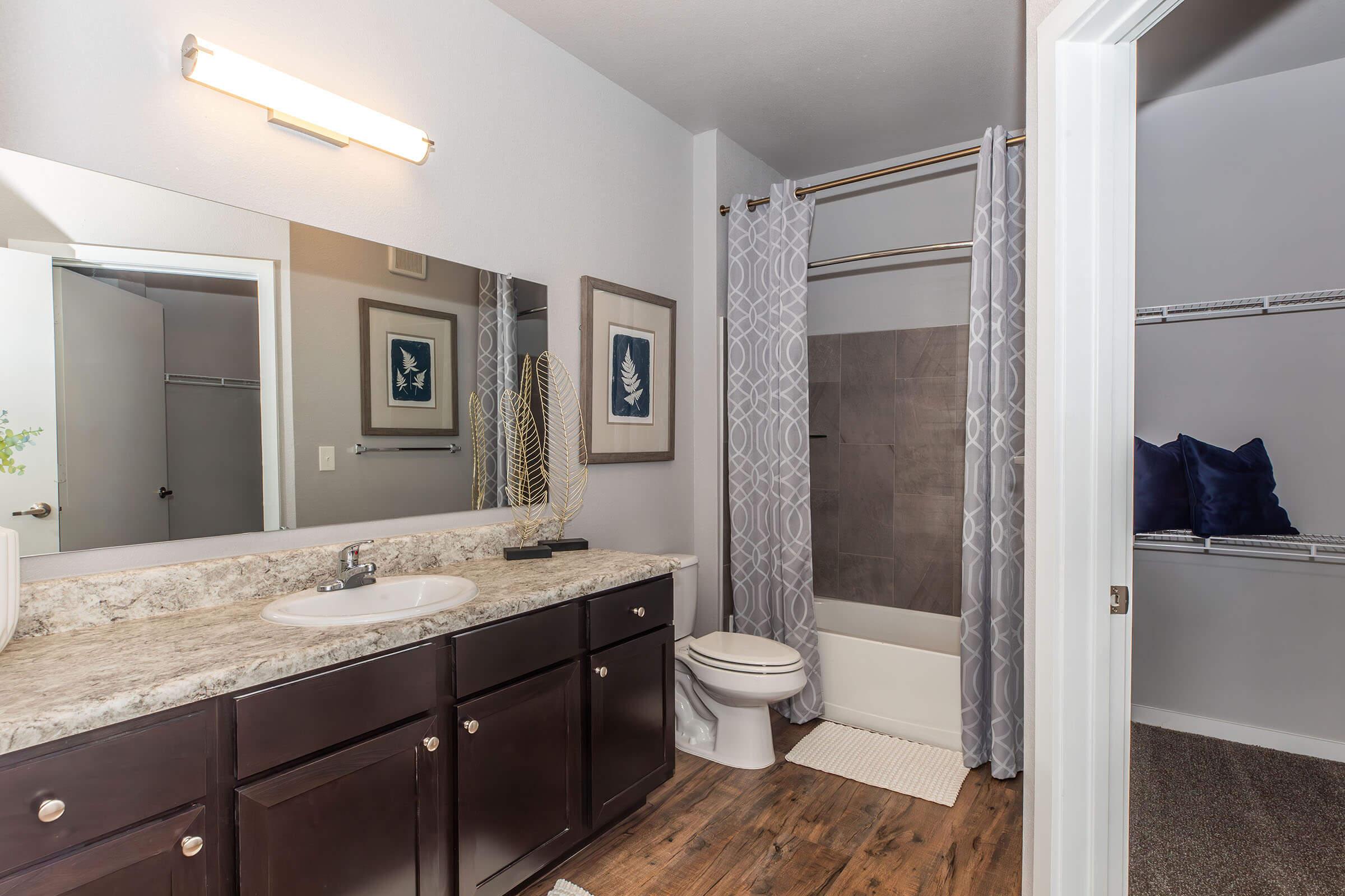 A modern bathroom featuring a dark wood vanity with a sink and large mirror, a white toilet, and a tub shower combination with gray tiles. The shower is enclosed with a textured curtain. Light gray walls complement the space, and a walk-in closet is visible on the right, showcasing shelves and navy blue pillows.