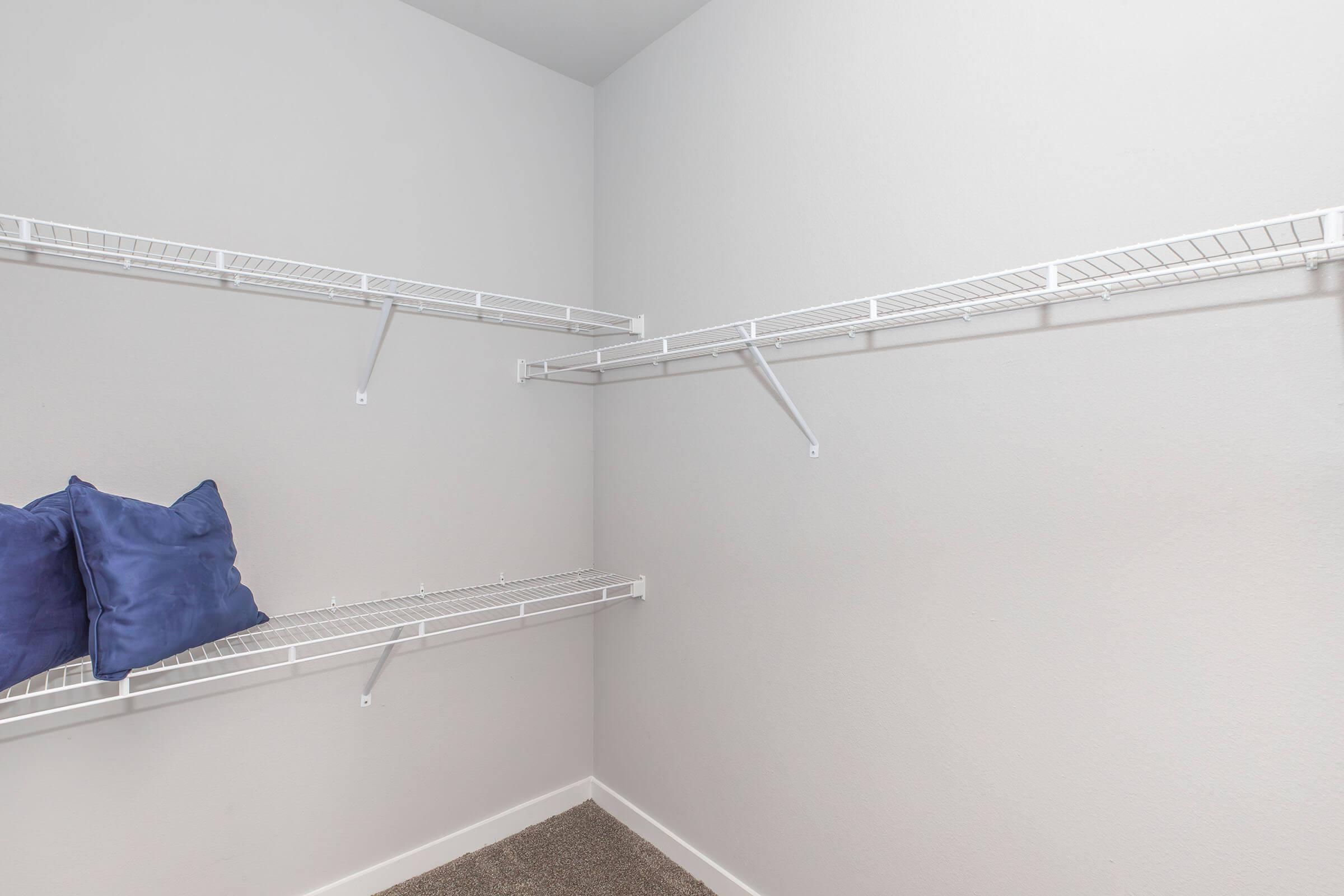Empty closet with white wire shelving on the walls. Two blue pillows are placed on the floor. The walls are painted a light grey, and the flooring appears to be carpeted.
