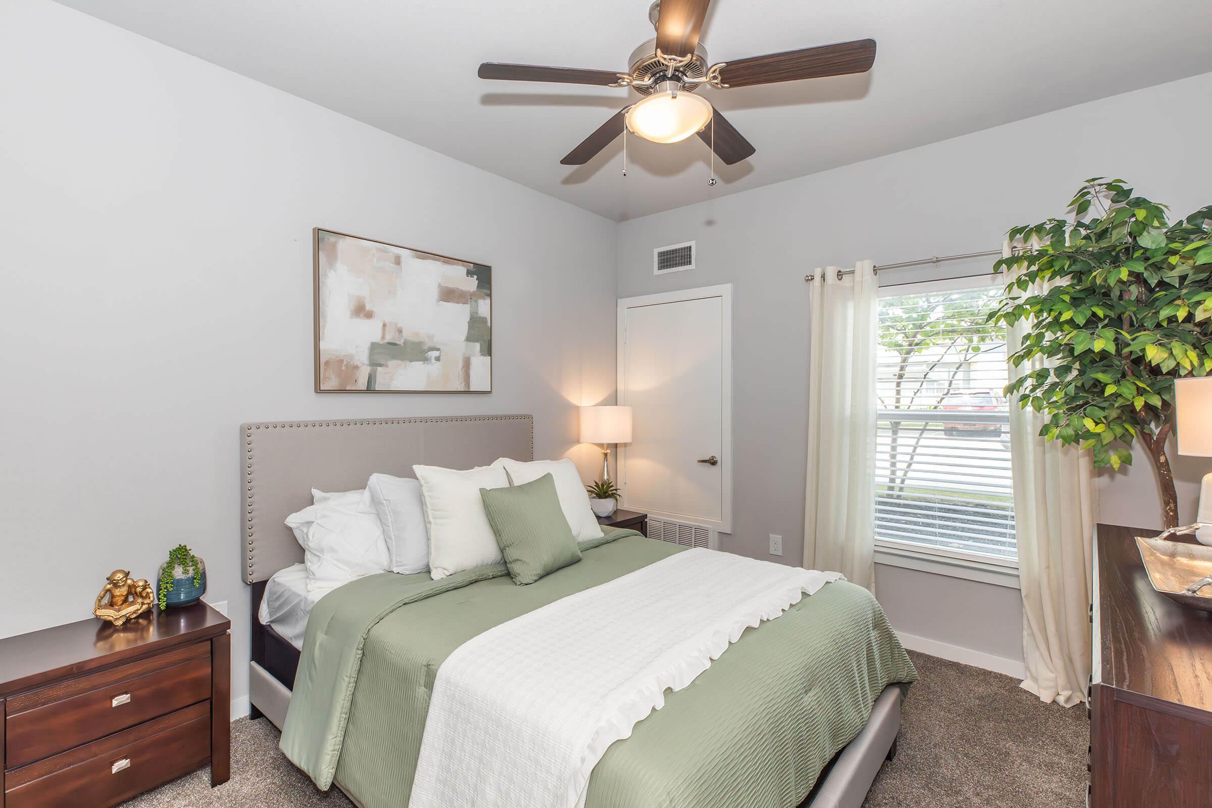 A well-decorated bedroom featuring a queen-sized bed with green and white bedding, a nightstand with a lamp, a window with light curtains, and a potted plant. The walls are painted light gray, and there is a fan on the ceiling along with modern artwork on the wall.