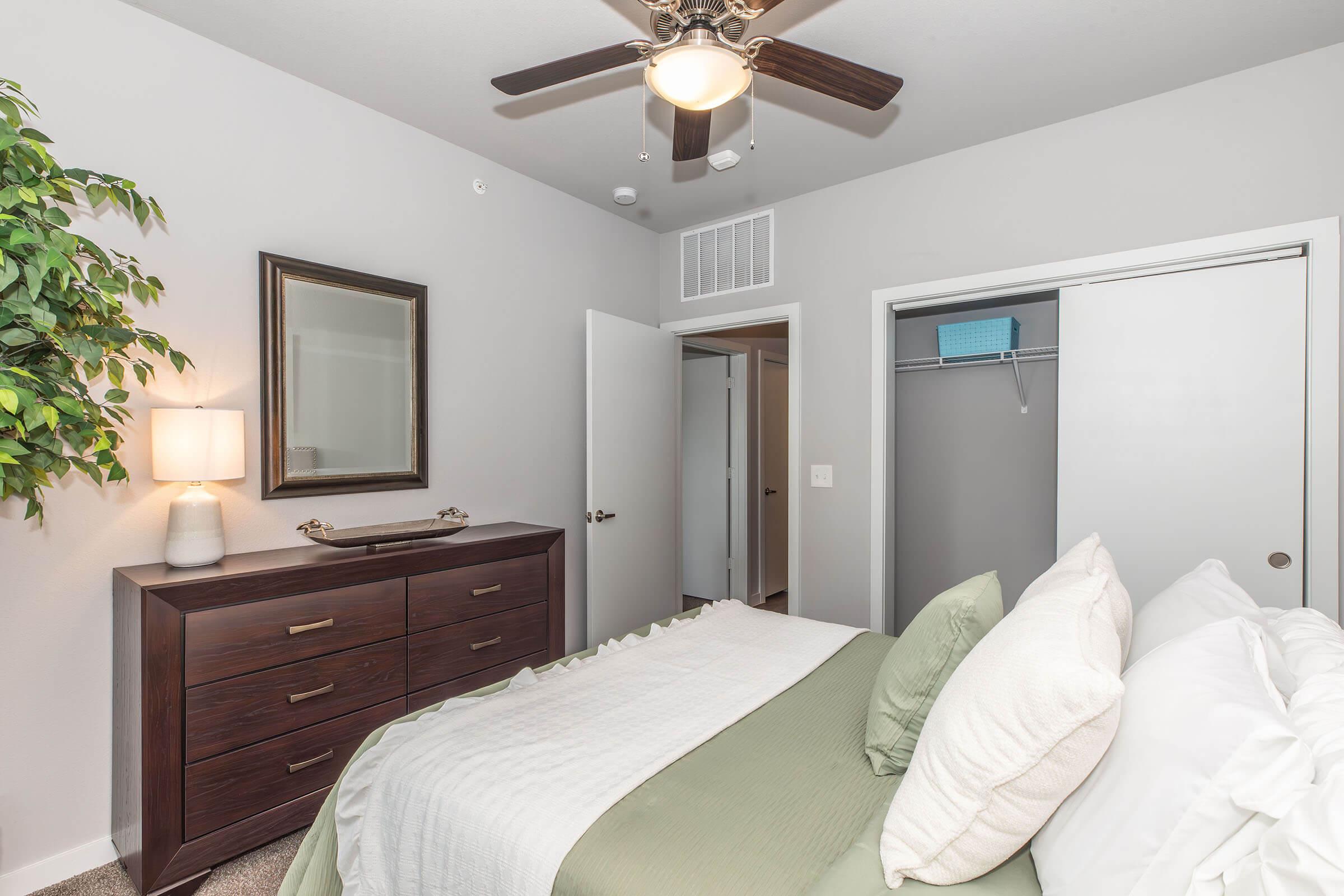 A cozy bedroom featuring a queen-sized bed with light green bedding, a dark wooden dresser, a lamp, and a decorative plant. There is a mirror above the dresser, a ceiling fan, and two doors leading to a closet and hallway. The walls are painted light gray, creating a calming atmosphere.