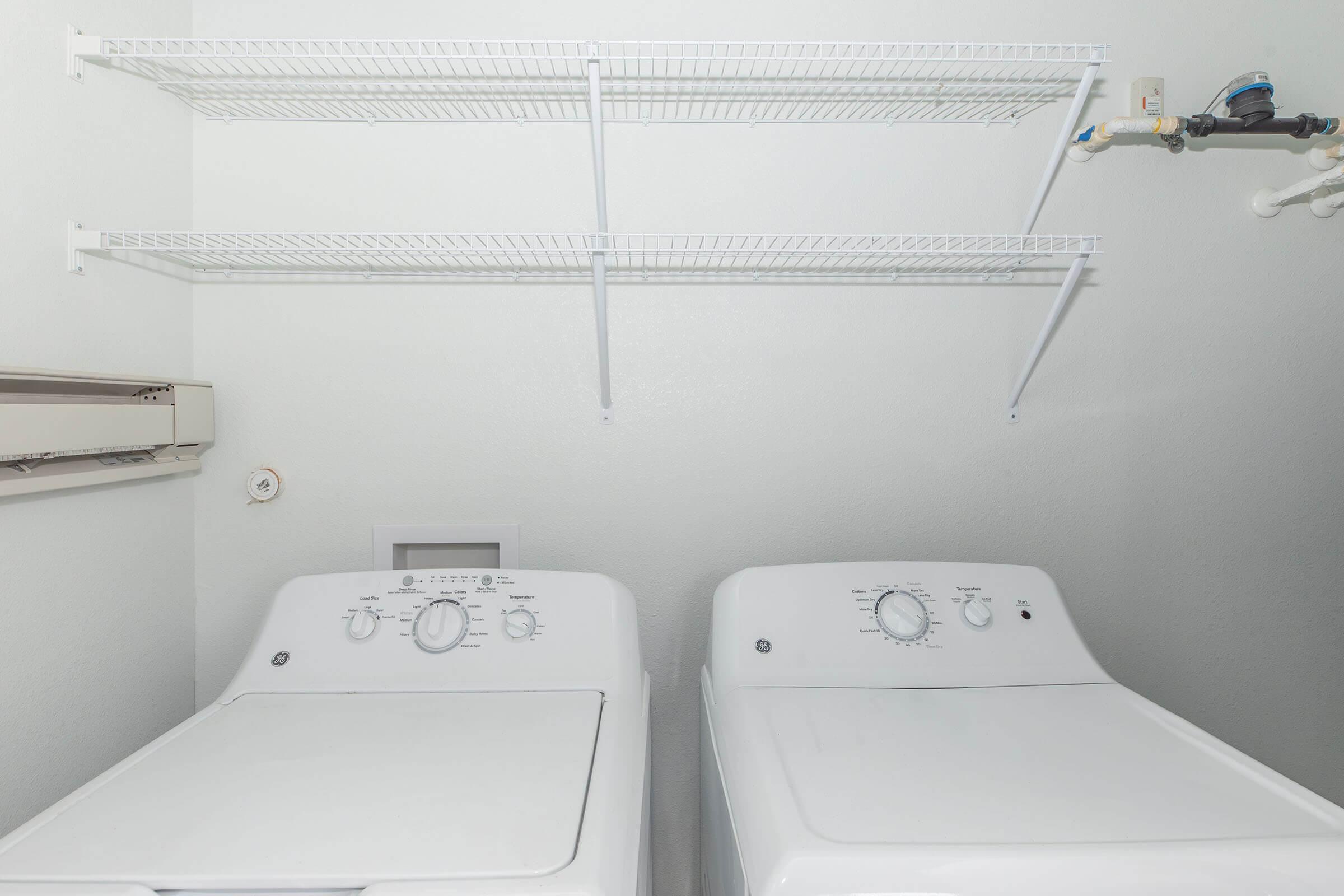 A clean laundry room displaying a white washing machine and dryer side by side. Above them, there are two white wire shelving units for storage. The walls are light-colored, creating a bright and organized space designed for household laundry tasks.