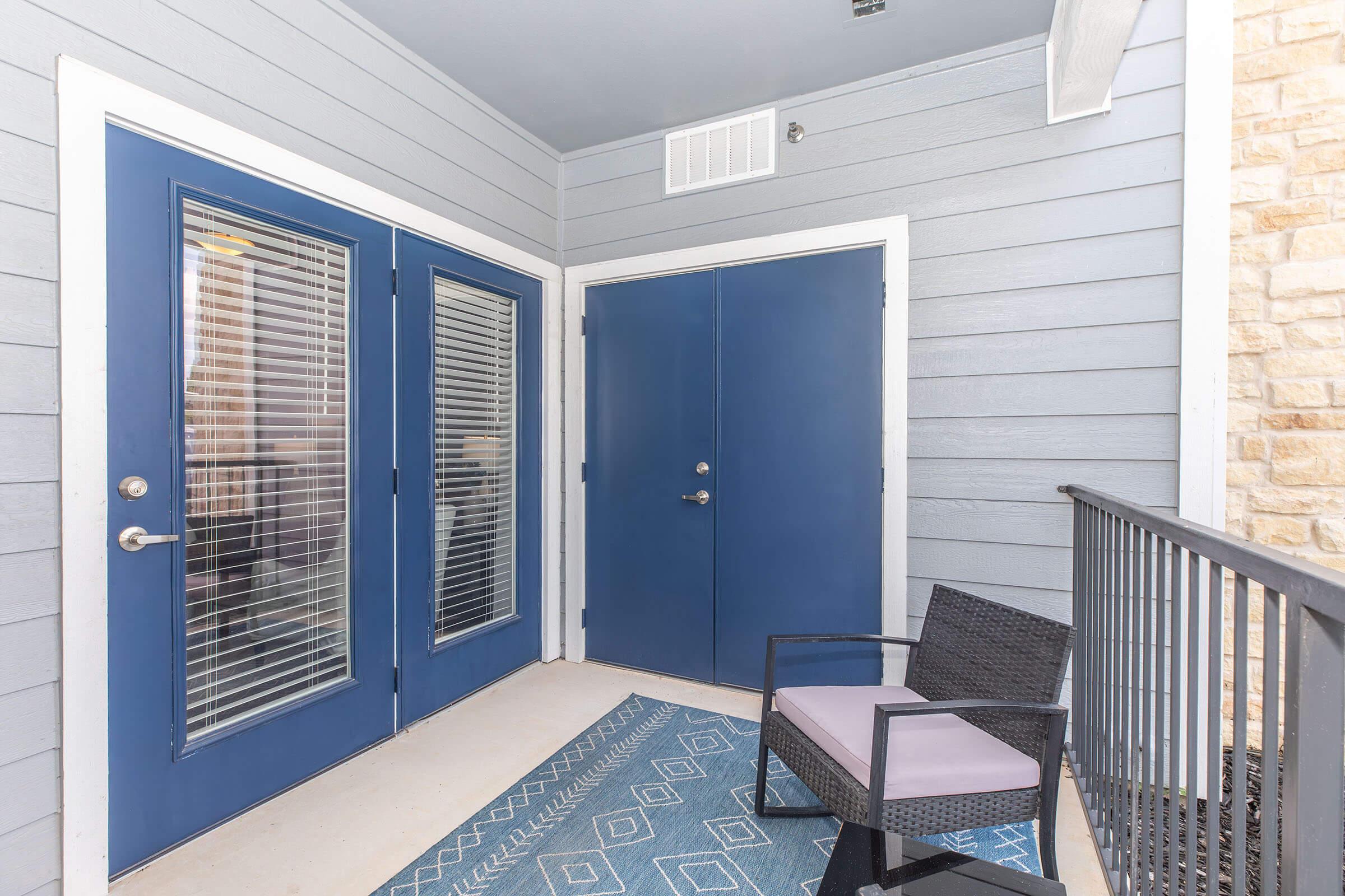 A welcoming exterior entryway featuring two blue doors with glass panels, a light blue wall finish, and a decorative area rug. A black wicker chair with a purple cushion is positioned next to the doors, enhancing the inviting atmosphere of the space.