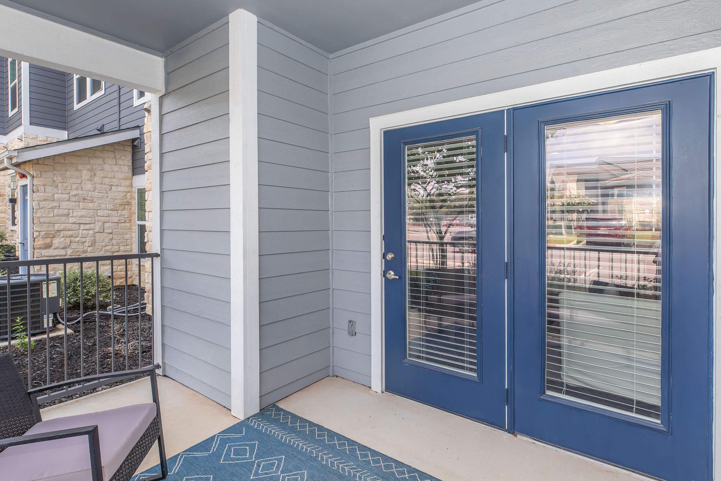 A blue front door with glass panels stands on a light gray porch, featuring a small seating area with a dark chair and a patterned rug. The entrance is framed by beige stone and gray siding, with a view of neighboring buildings visible through the doors.