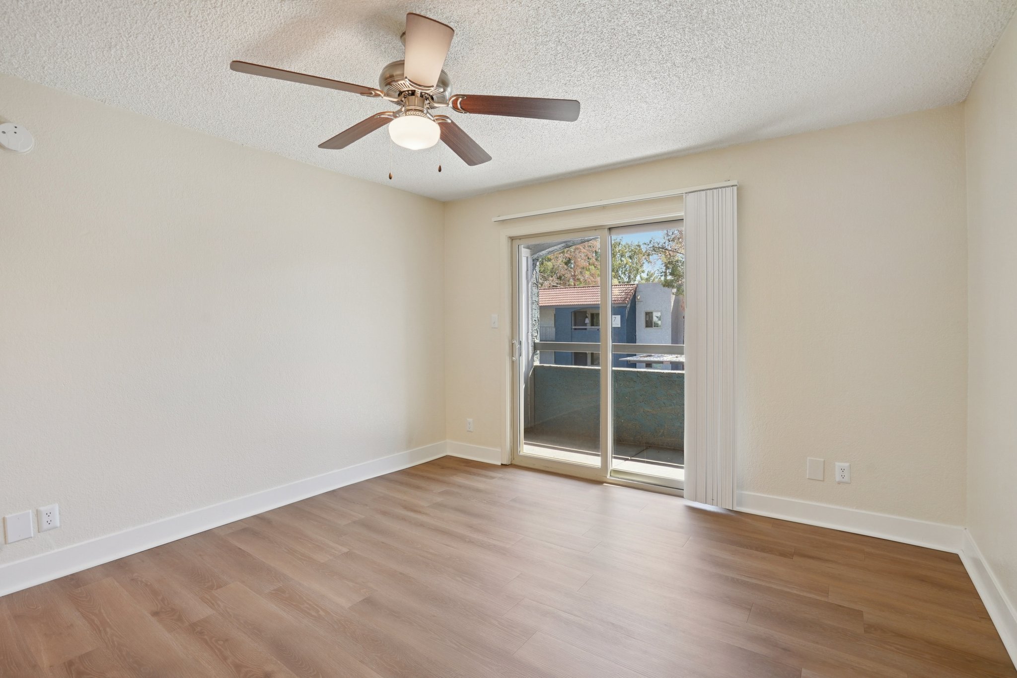 Empty room with light-colored walls and a ceiling fan. It features a sliding glass door leading to a small balcony, and large windows allow natural light. The flooring is a warm-tone laminate, creating a cozy atmosphere.