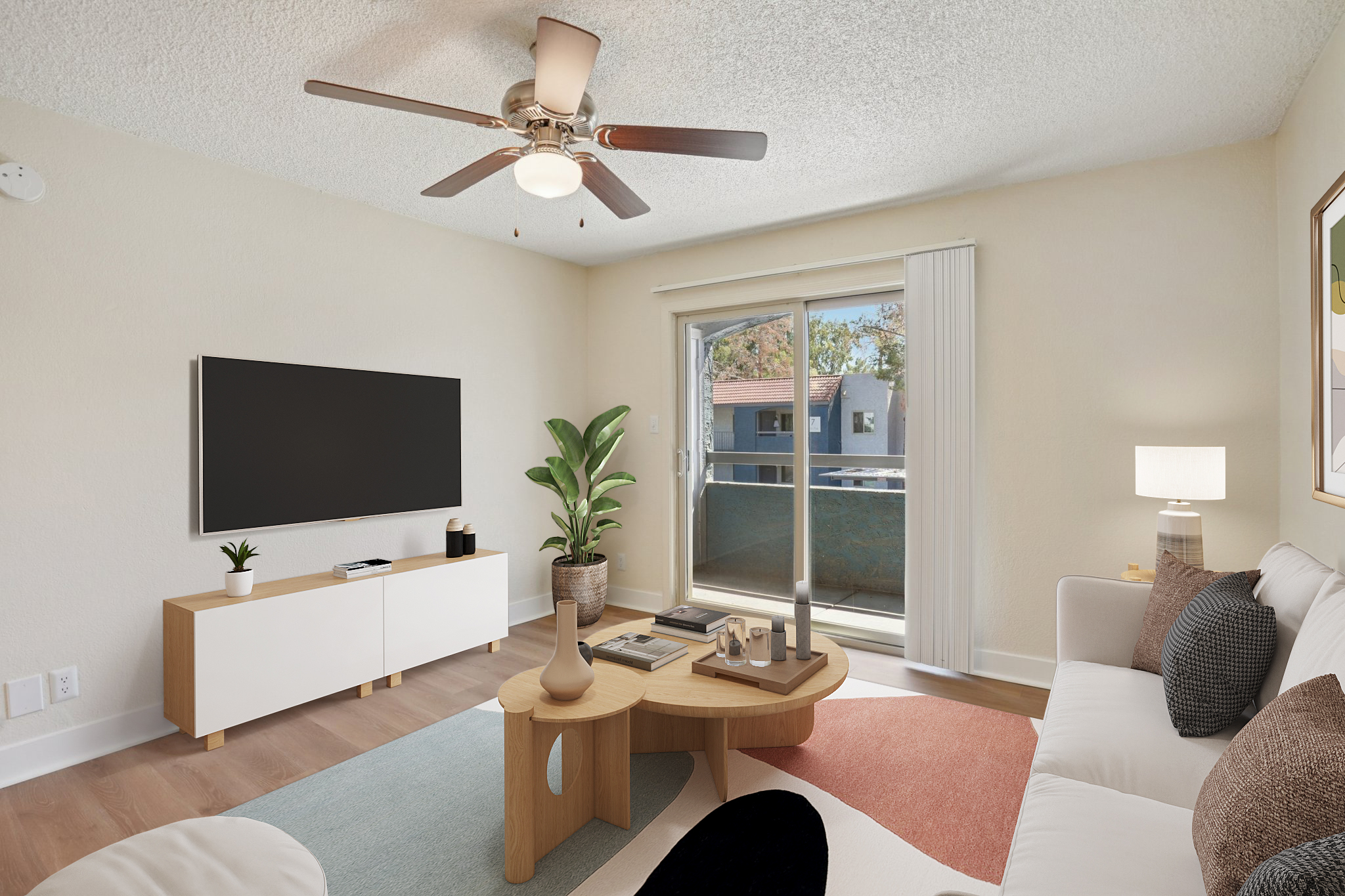 A modern living room with a white sofa, a wooden coffee table, and a large flat-screen TV mounted on the wall. There's a potted plant beside the coffee table and a lamp on a side table. A sliding glass door leads to a balcony, with light-colored walls and a pastel area rug adding warmth to the space.