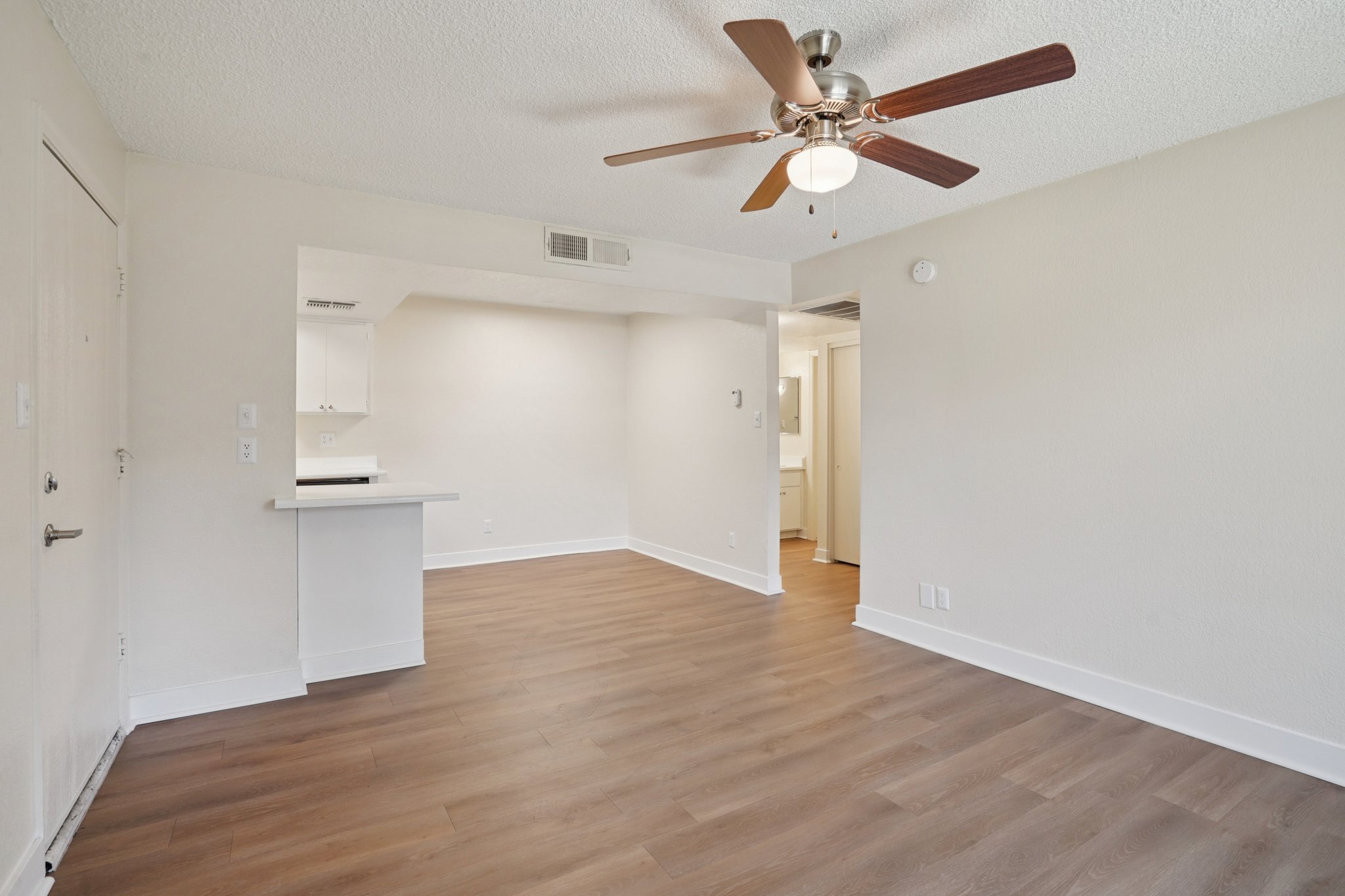 An interior view of a bright and spacious living area featuring a ceiling fan, light-colored walls, and hardwood flooring. The layout includes an open area that connects to a kitchen space visible in the background, creating a warm and inviting atmosphere.