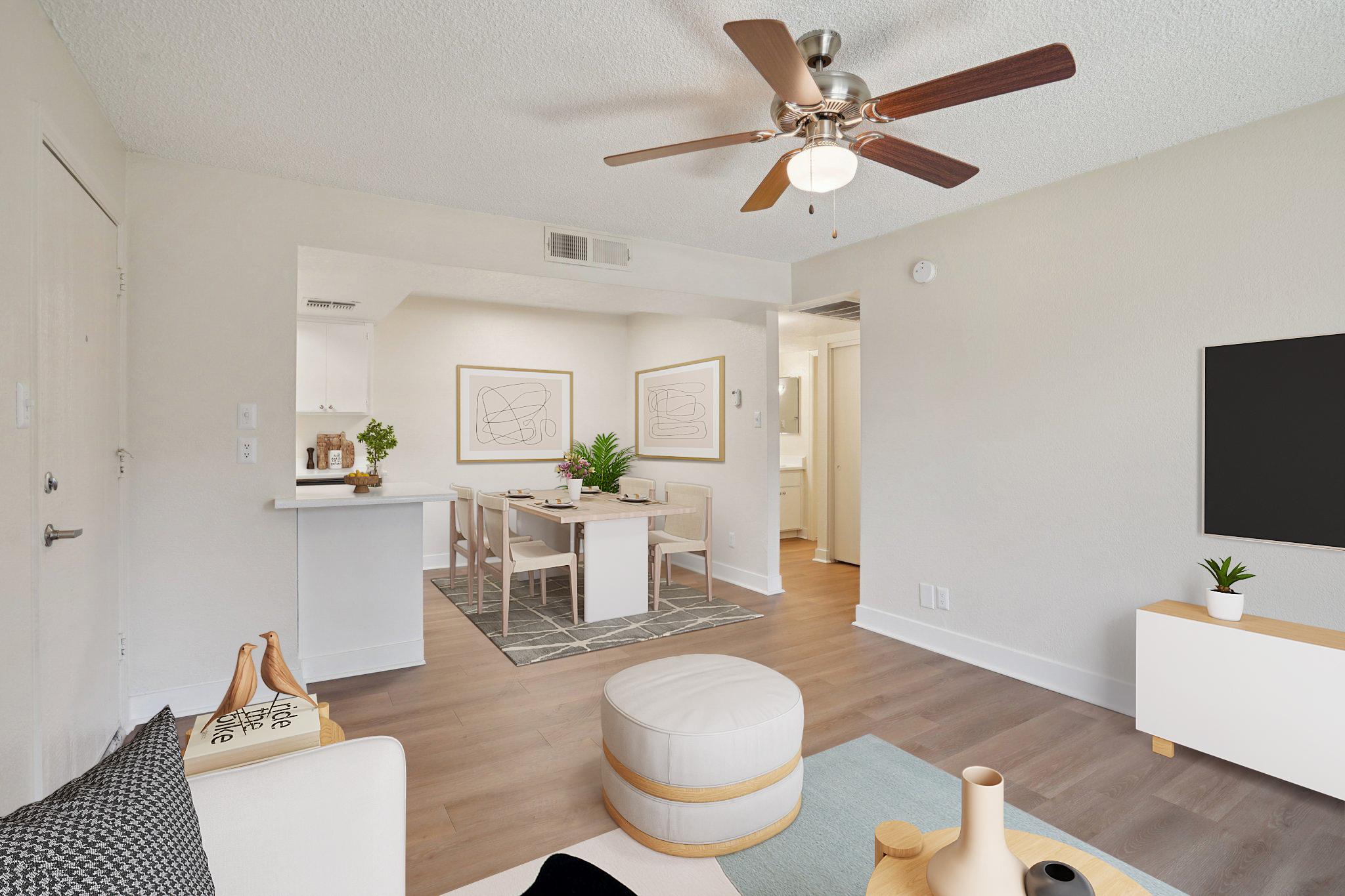 A bright, modern living room featuring a ceiling fan, neutral walls, and hardwood flooring. A white coffee table and pouf are in the foreground, while a dining area with a white table and chairs is visible in the background. Framed minimalist art and plants add decorative touches.