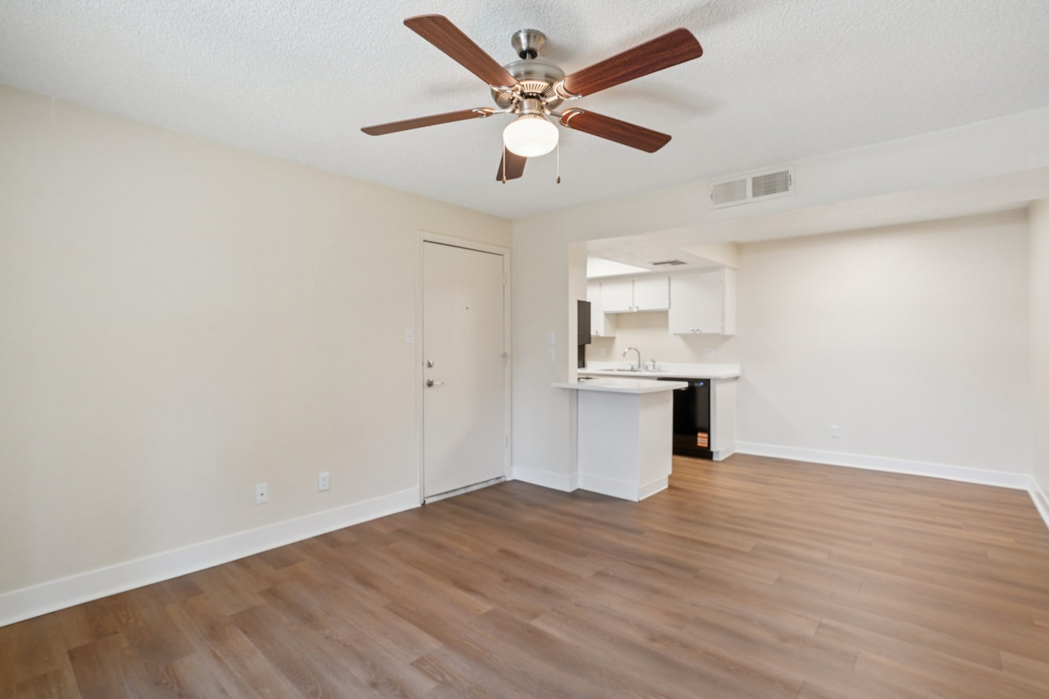 Interior view of an apartment with light-colored walls, a ceiling fan with wooden blades, and a spacious layout. The floor is made of wooden planks, and there’s an open area leading to a kitchenette in the background. Natural light fills the space, creating a bright and inviting atmosphere.