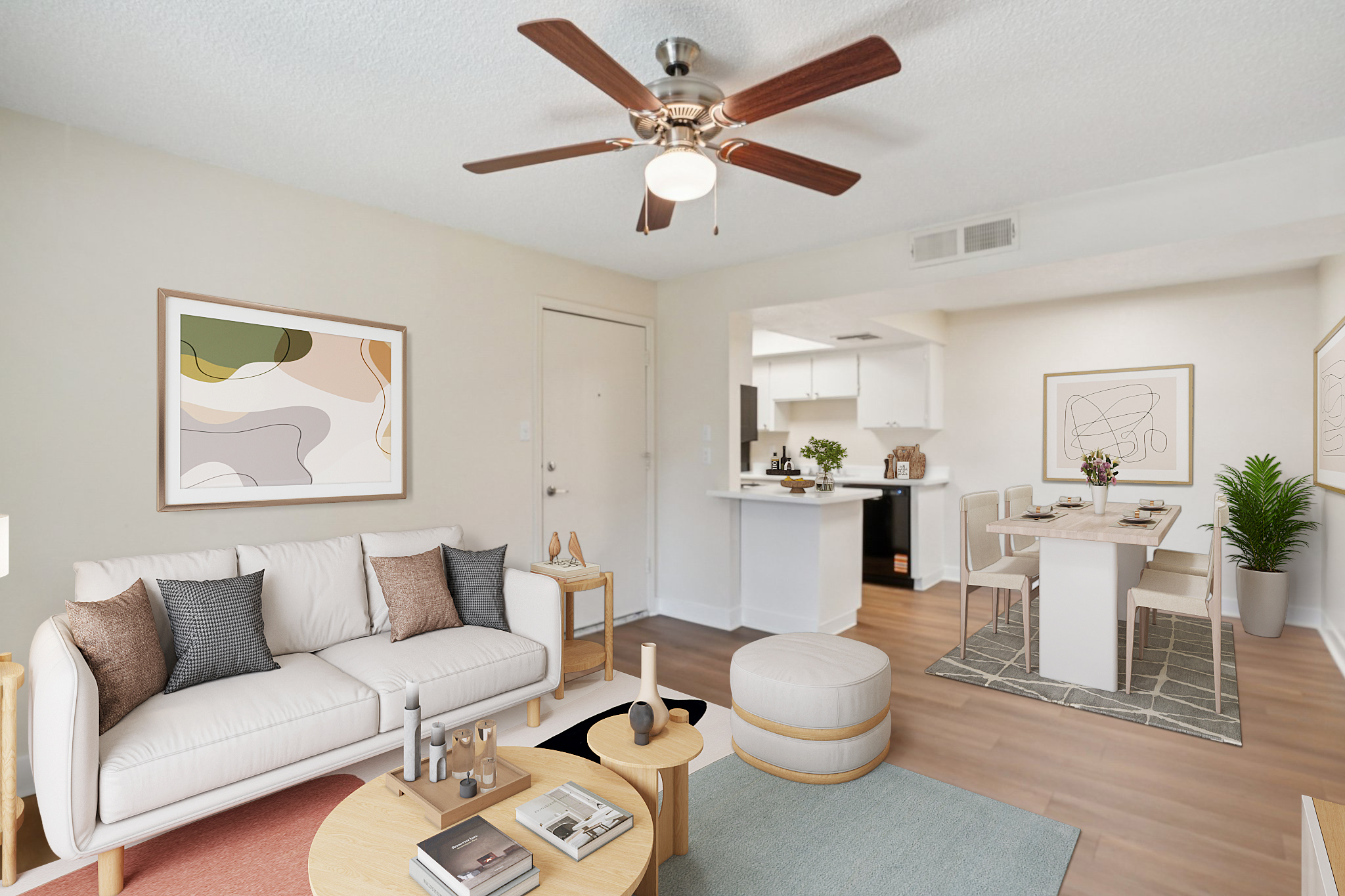 A modern living room featuring a light-colored sofa with decorative pillows, a wooden coffee table, and a round ottoman. There is a ceiling fan, framed abstract art on the wall, and a dining area with a table set for two. The space has a neutral color scheme and warm lighting, suggesting a cozy atmosphere.
