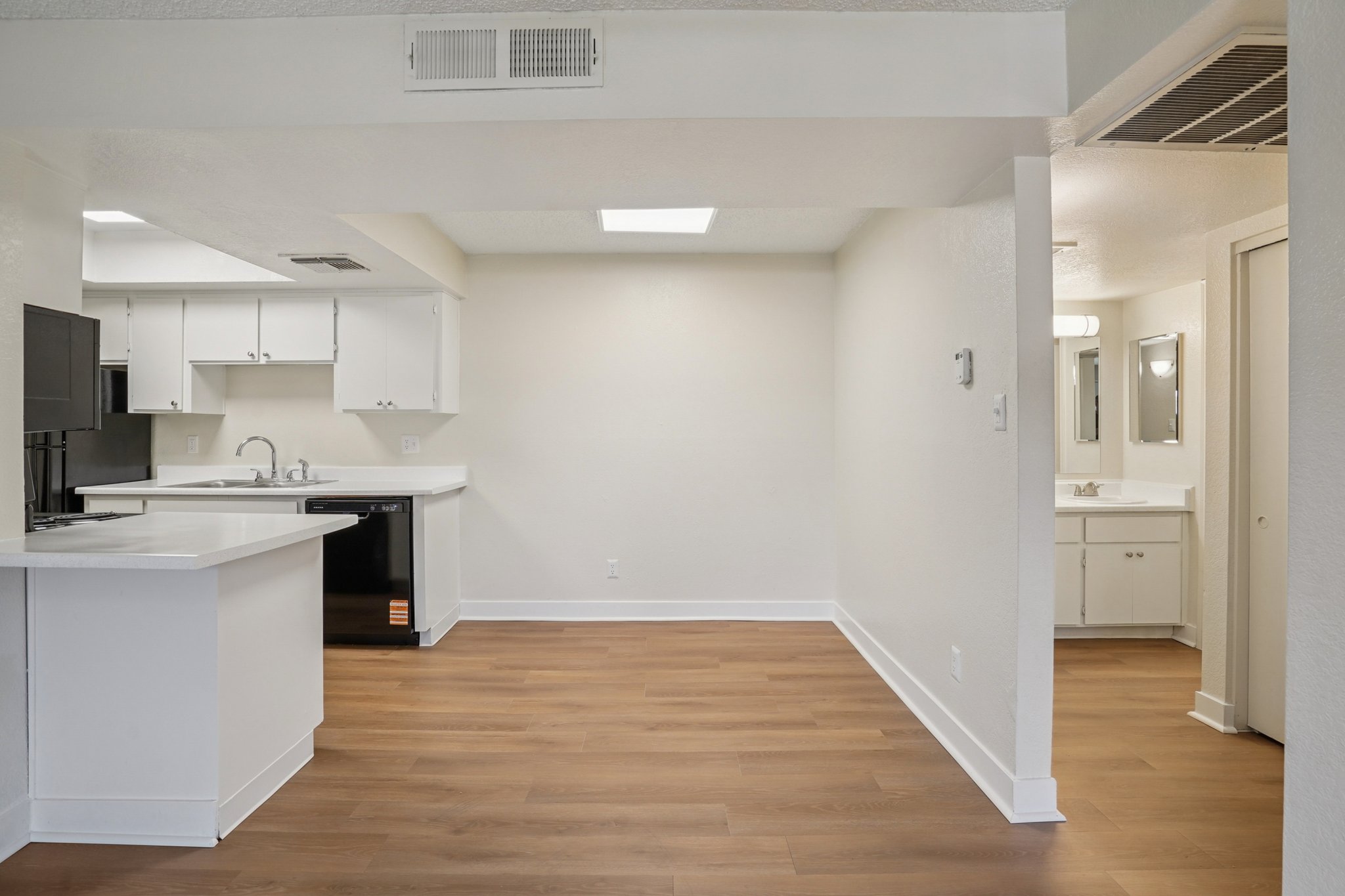 A bright, modern open-concept living area with light wood flooring. The kitchen features white cabinets and black appliances, with a countertop separating it from the living space. The area is well-lit with natural light, and there are two doors leading to additional rooms or areas.