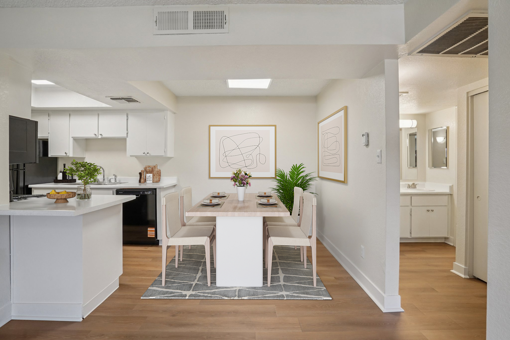 A modern dining area featuring a white table with six chairs, set for a meal with plates and floral centerpieces. The open kitchen is visible in the background, showcasing sleek appliances and a clean design. Light-colored walls and wooden flooring create a bright, inviting atmosphere, complemented by abstract wall art.