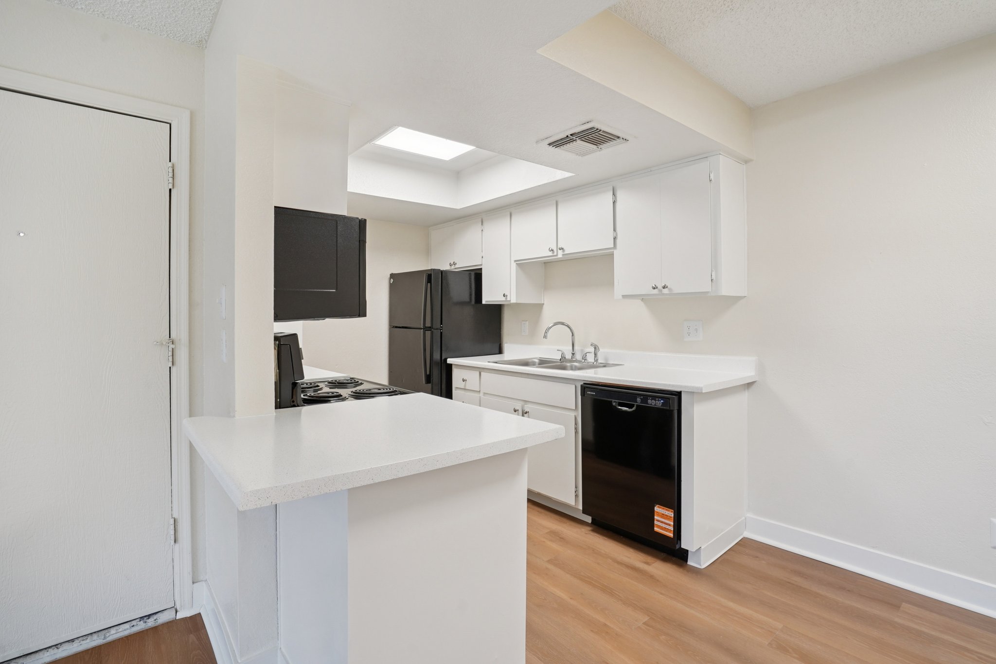 Modern kitchen featuring white cabinets, a black refrigerator, and stainless steel appliances. The space includes a counter with bar seating, a skylight for natural light, and wood-style flooring, creating a bright and inviting atmosphere.