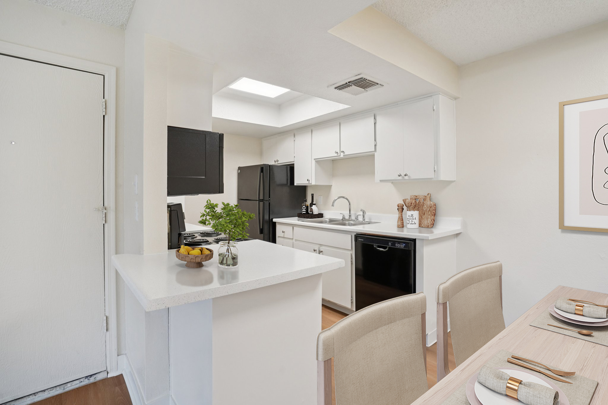 Modern kitchen featuring white cabinetry, black appliances, and a granite countertop. There is a small dining area with a wooden table set for four, and a bowl of fruit on the counter. Natural light enters through a skylight, creating a bright atmosphere in the space.