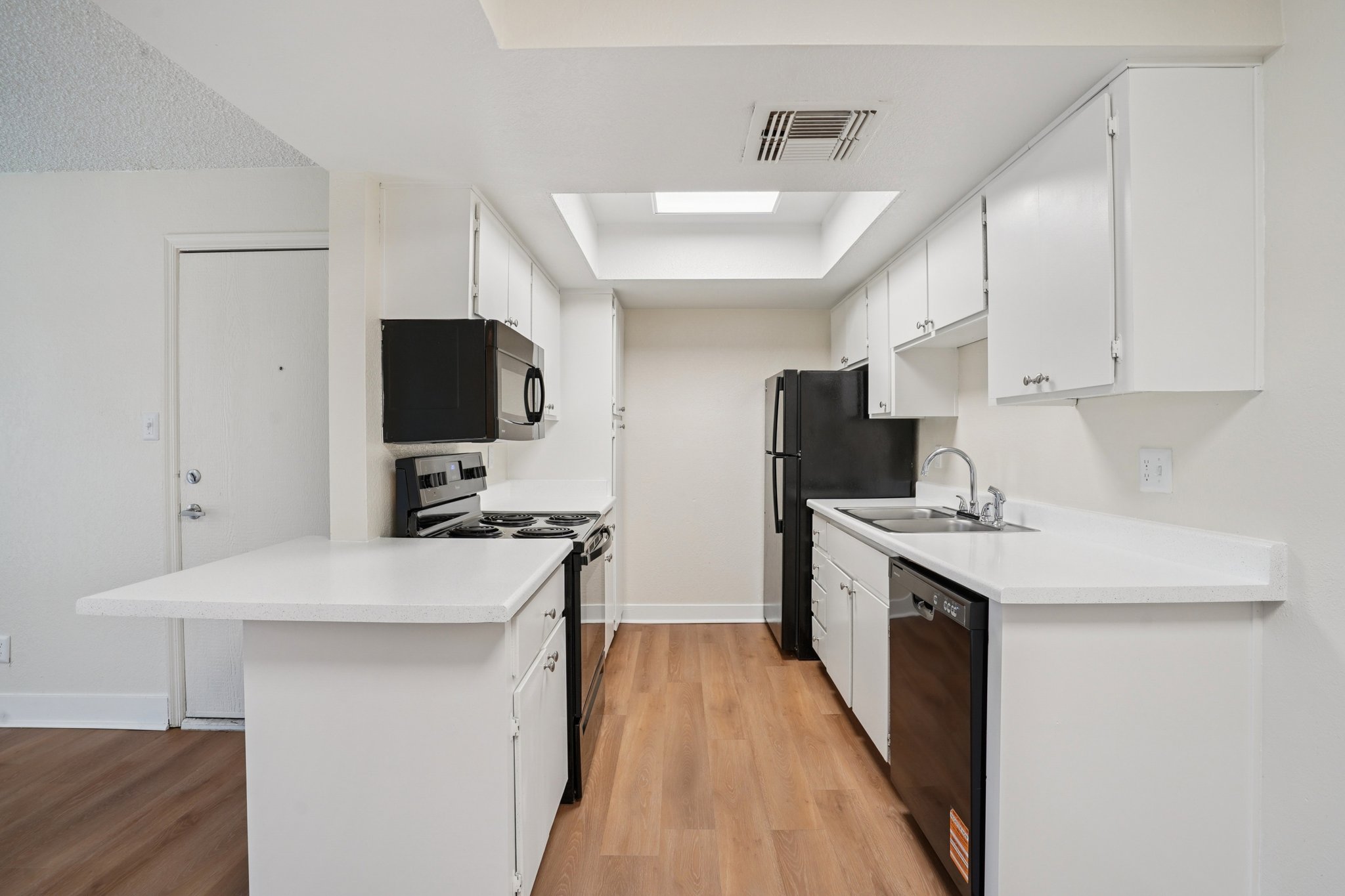 Modern kitchen featuring white cabinets, stainless steel appliances, and a spacious layout. Includes a double sink, black refrigerator, microwave, and stove. Natural light from a skylight brightens the area. Hardwood floors create a warm atmosphere. The kitchen island offers additional counter space.