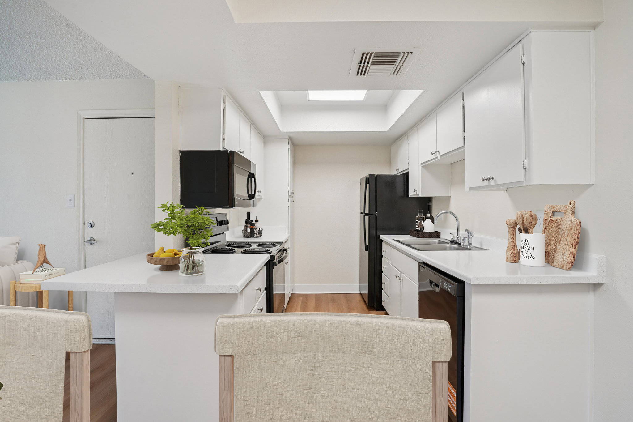 A bright and modern kitchen featuring white cabinets, stainless steel appliances, and a central island. Natural light streams in from a skylight above, enhancing the open feel. Decorative items, including a plant and a bowl of fruit, add a touch of warmth to the space.