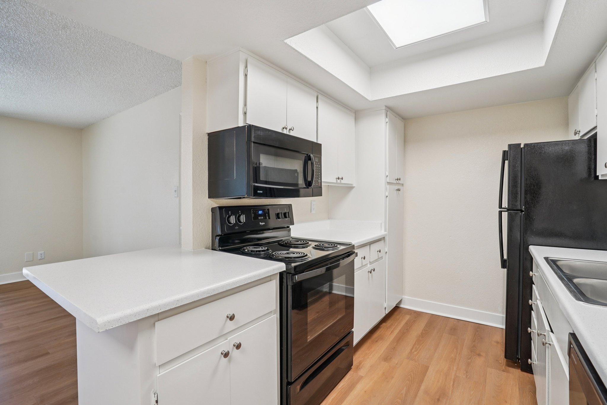 A modern kitchen featuring white cabinets, a black stove and microwave, a black refrigerator, and a light-colored countertop. The kitchen has a skylight overhead, providing natural light, and is designed with an open layout leading into a living space with neutral walls and wooden flooring.