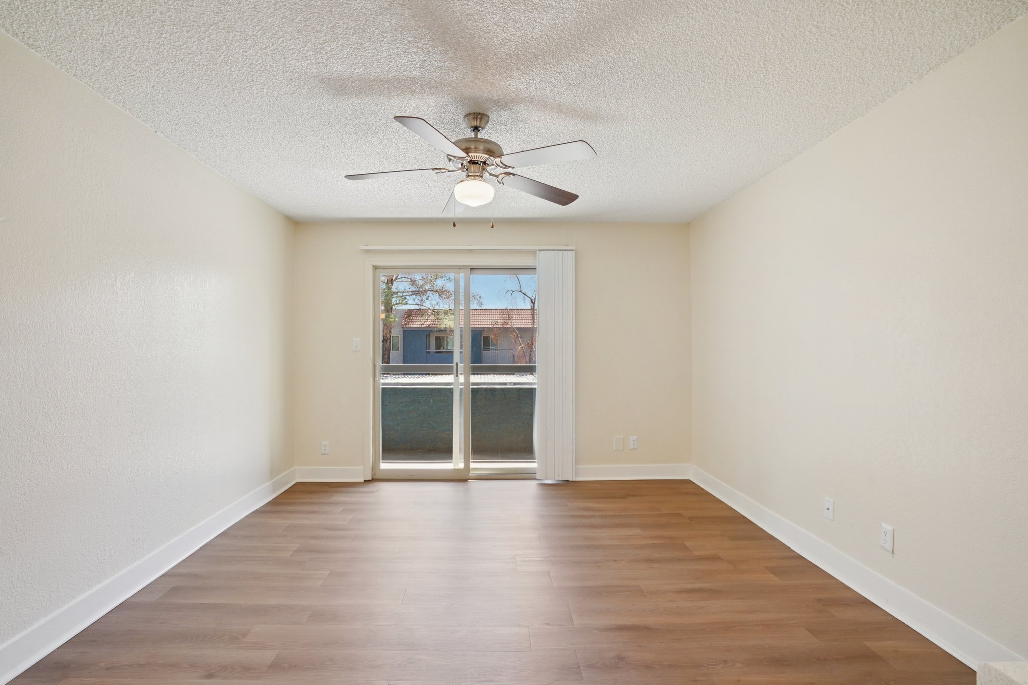 Spacious empty living room with a ceiling fan and sliding glass doors leading to a balcony. The room features light-colored walls and a wood-look floor. Natural light illuminates the space, creating an inviting atmosphere. Suitable for various furniture arrangements.