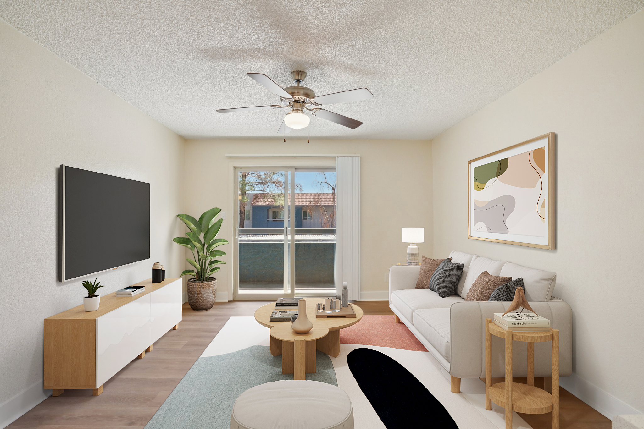 A modern living room featuring a light-colored sofa, a circular coffee table, and a TV on a white cabinet. A large window brings in natural light, with a potted plant and an abstract art piece on the wall. The floor has a colorful area rug, and there's a ceiling fan above.