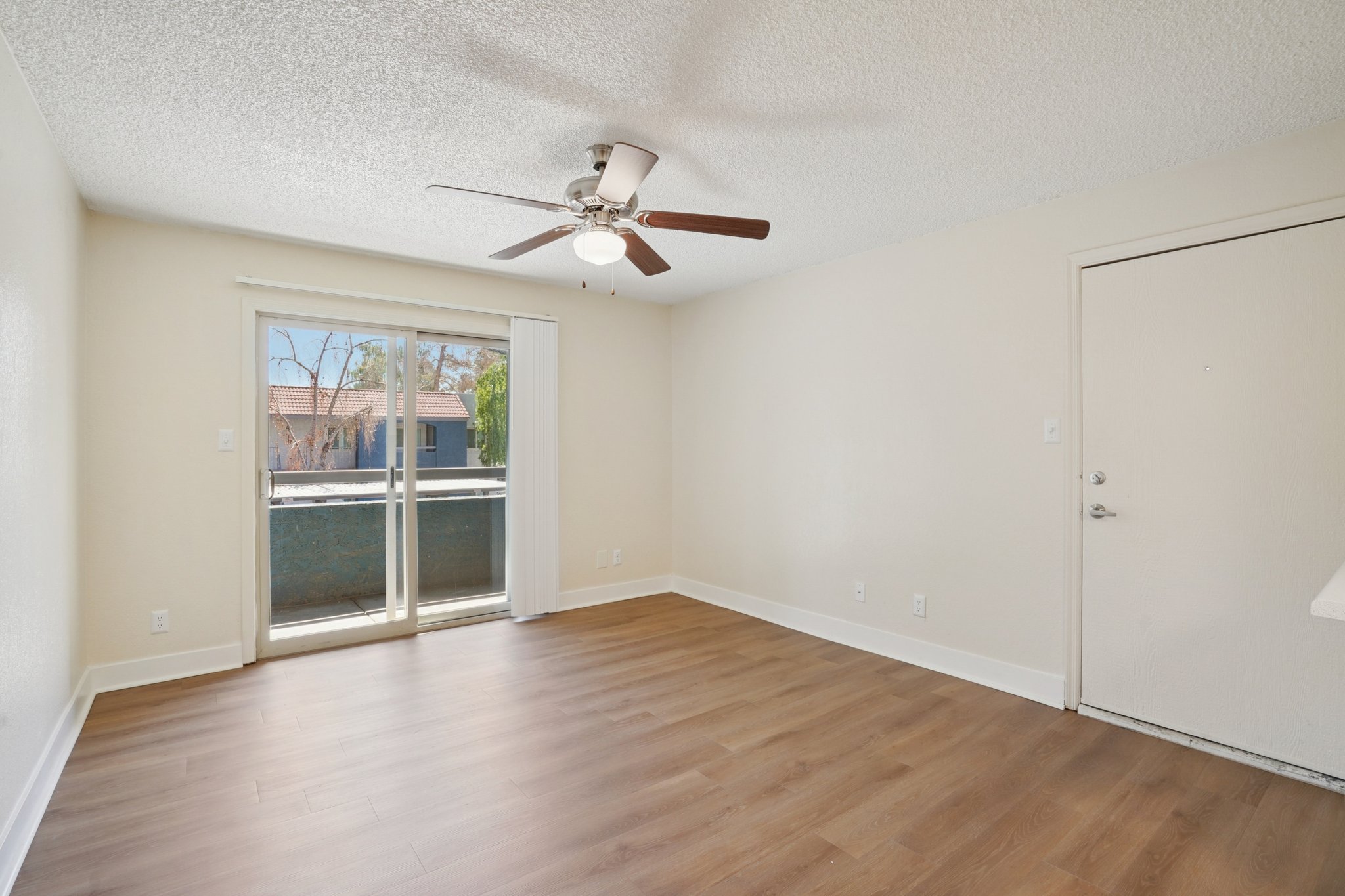 Empty room with light-colored walls, a ceiling fan, and a sliding glass door leading to a small balcony. The flooring is wooden, and there is a door on the right side. Natural light enters through the glass door, enhancing the spacious feel of the area.