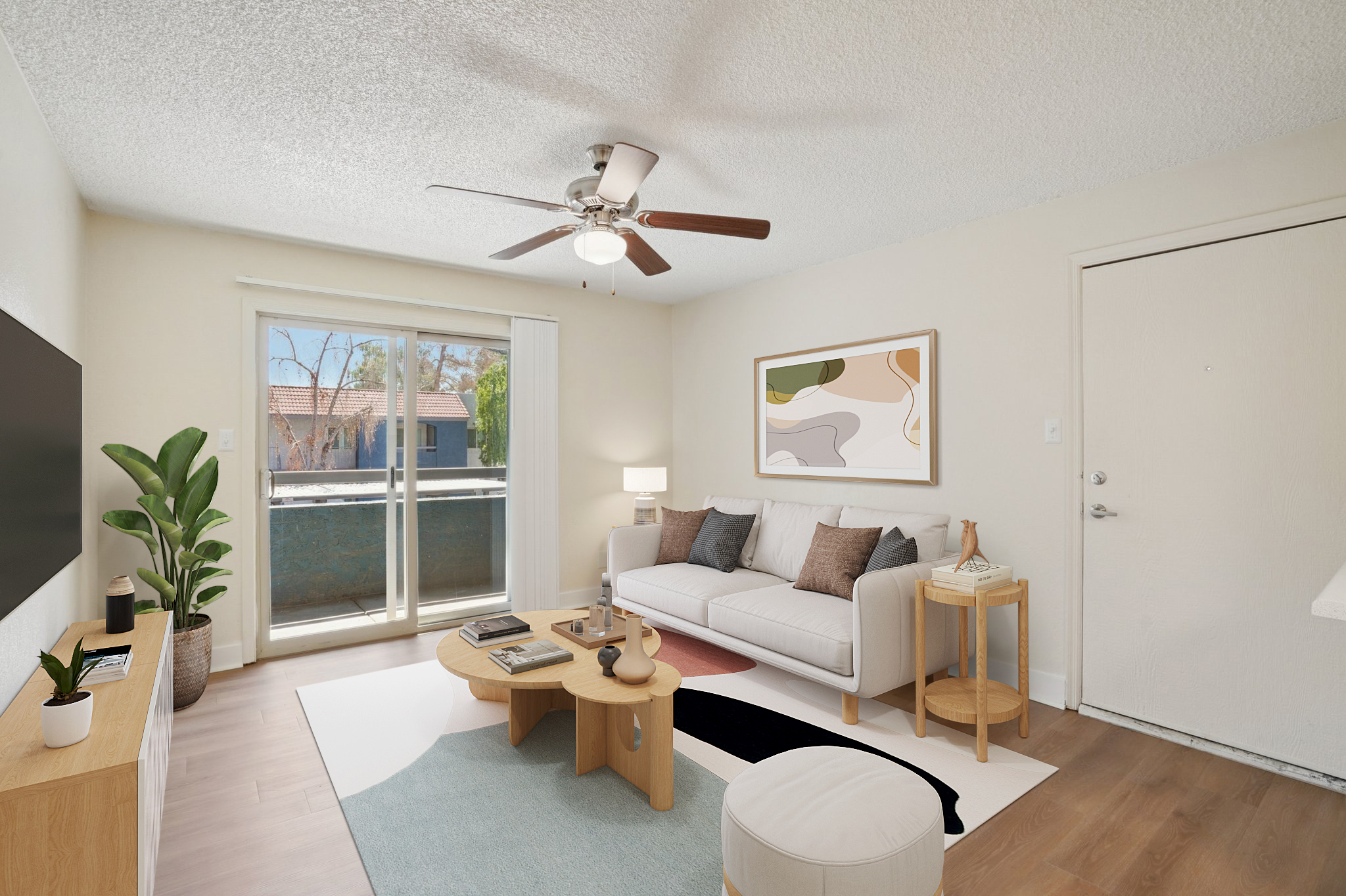 Bright and modern living room with a white sofa adorned with colorful cushions. A wooden coffee table sits on a patterned rug, surrounded by a leafy plant. A large window allows natural light, and a ceiling fan hangs above. The wall features an abstract art piece, and there's a door leading outside.