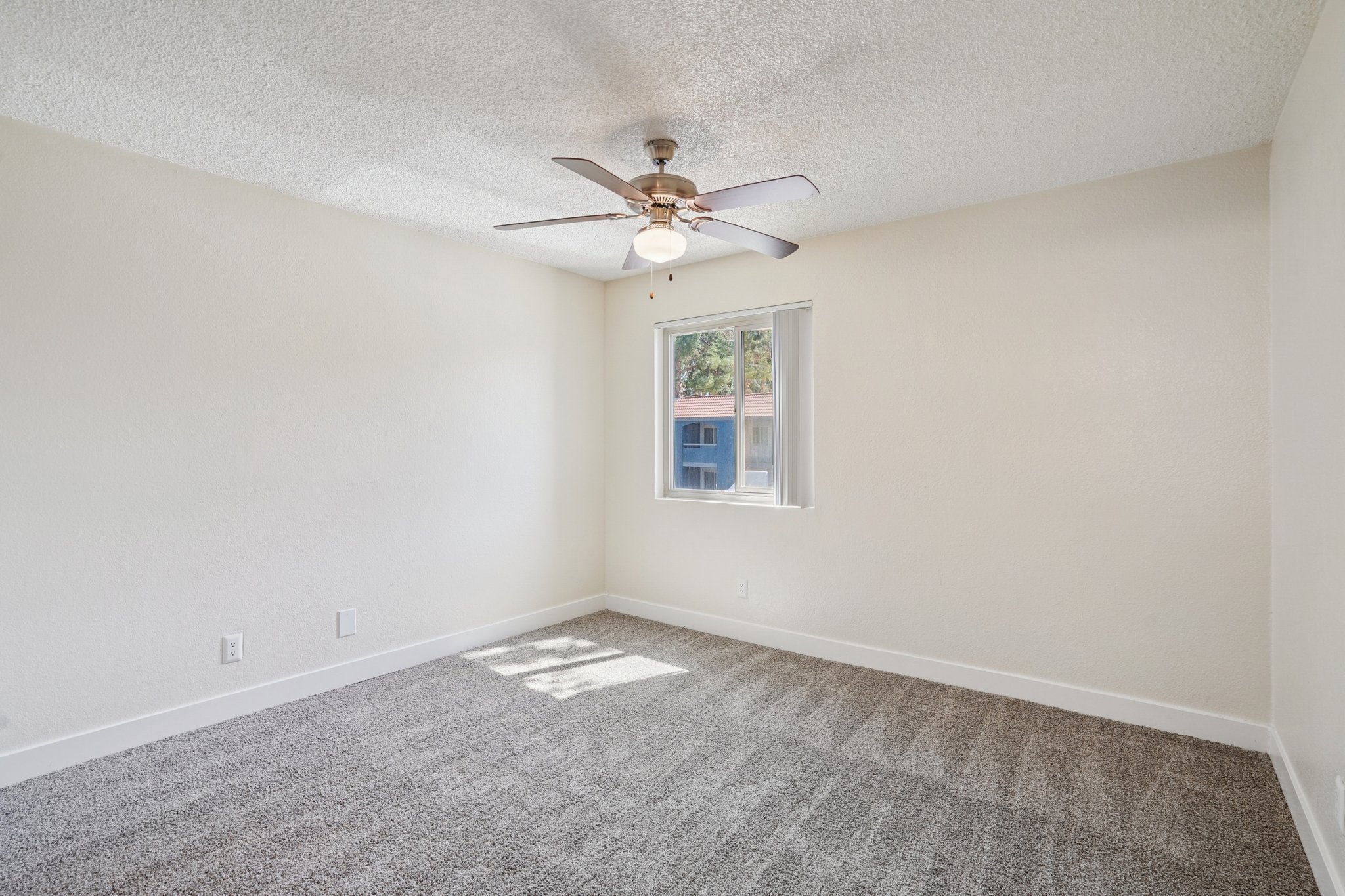 A spacious, well-lit room with light-colored walls and a ceiling fan. The floor is carpeted in a soft gray, and a window allows natural light to fill the space. The overall atmosphere is welcoming and ready for personal touches.
