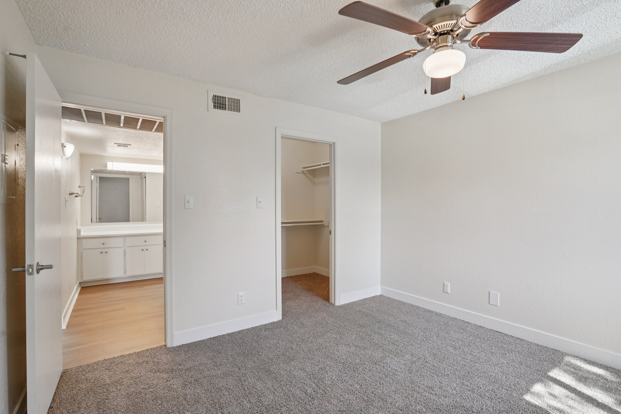 A well-lit, empty bedroom featuring beige walls and a light brown carpet. It includes a ceiling fan with a light fixture, a closet on the right, and an open doorway leading to a bathroom area with double sinks. The room has a clean, modern appearance.