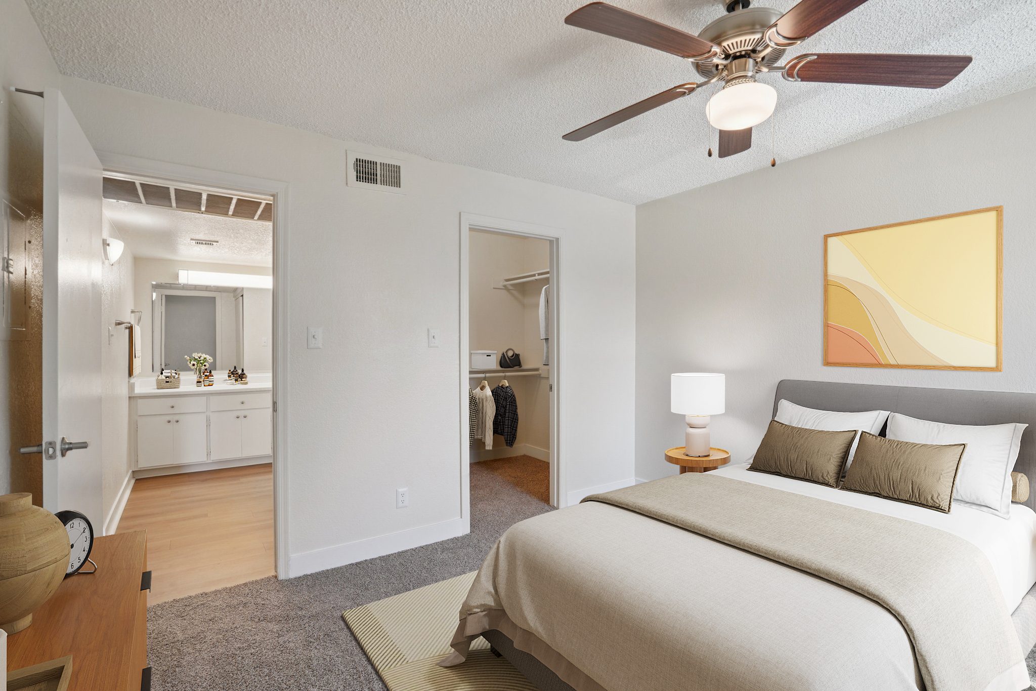 A cozy bedroom featuring a queen-sized bed with neutral-colored linens and decorative pillows. A ceiling fan hangs above, and a small bedside lamp is present. The room includes a doorway to a closet and a bathroom visible in the background, with natural light illuminating the space and a minimalist artwork on the wall.