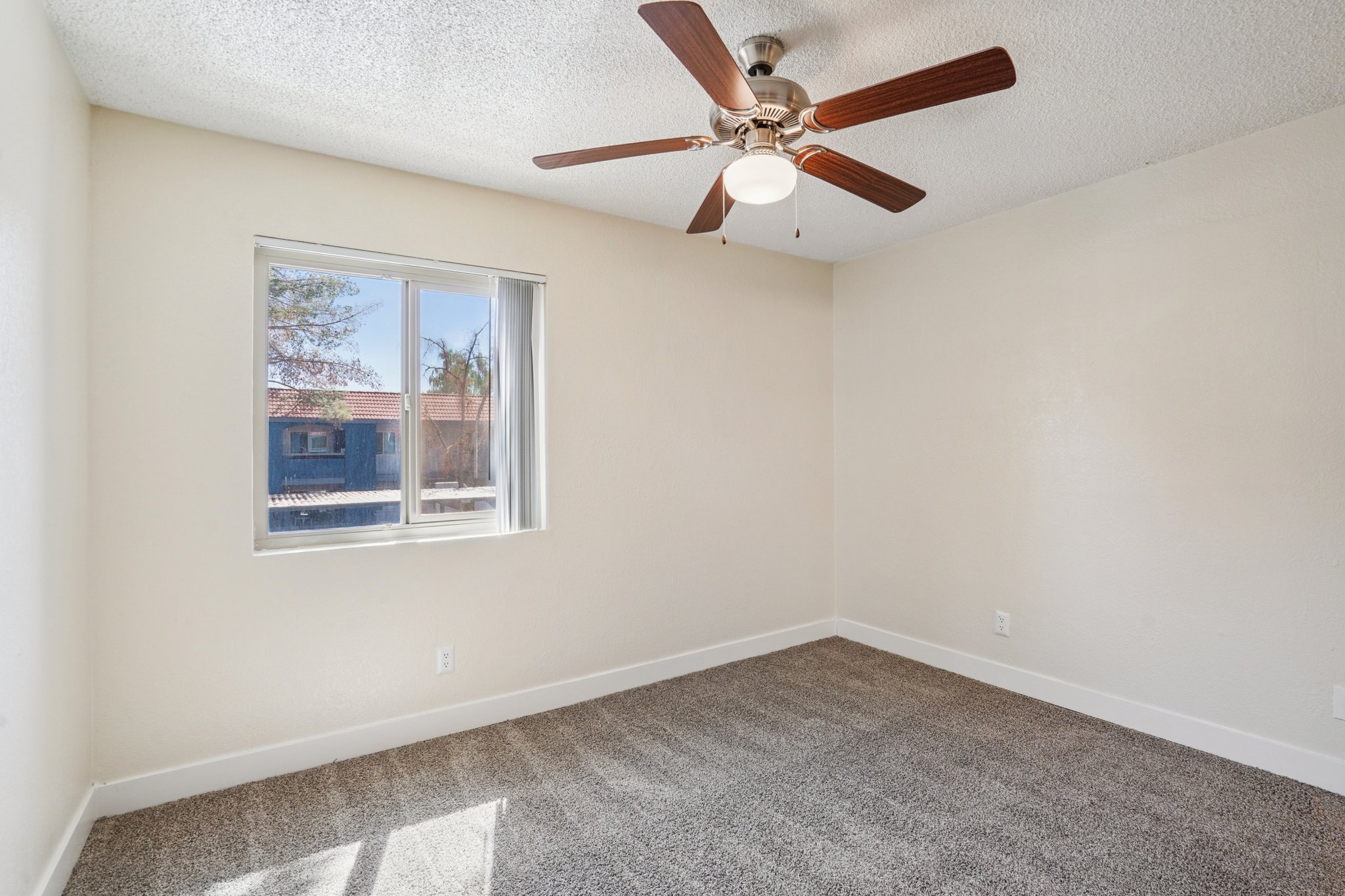 A spacious, empty room with light beige walls and a ceiling fan. Natural light streams in through a window, illuminating the soft gray carpet. The room features a simple design and a bright, clean atmosphere, ideal for various uses.