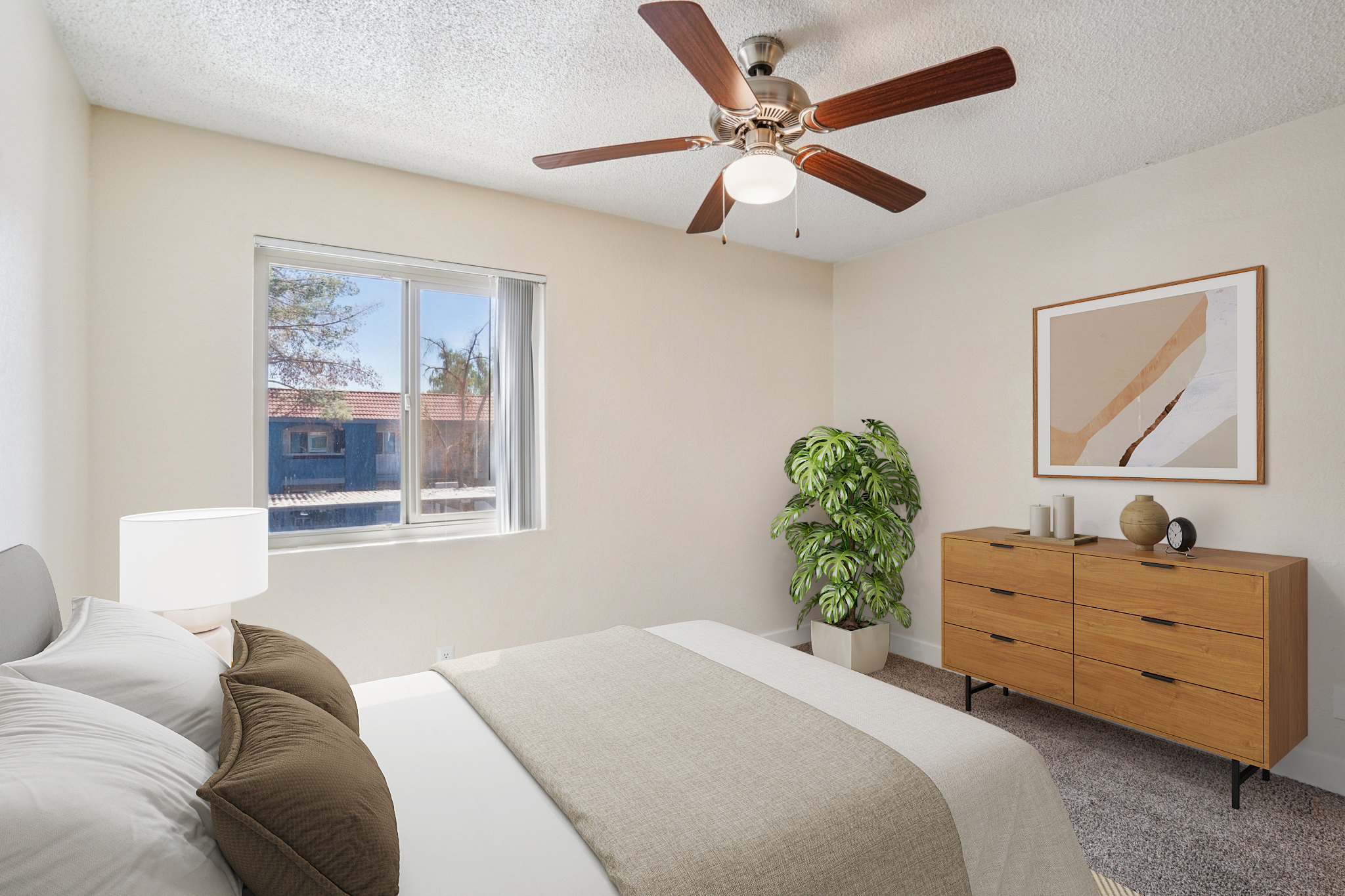 A cozy bedroom featuring a light-colored wall, a large window with natural light, a ceiling fan, and a neatly made bed with neutral bedding. A wooden dresser sits against one wall, accompanied by a decorative plant and artwork. The room exudes a calm and inviting atmosphere.