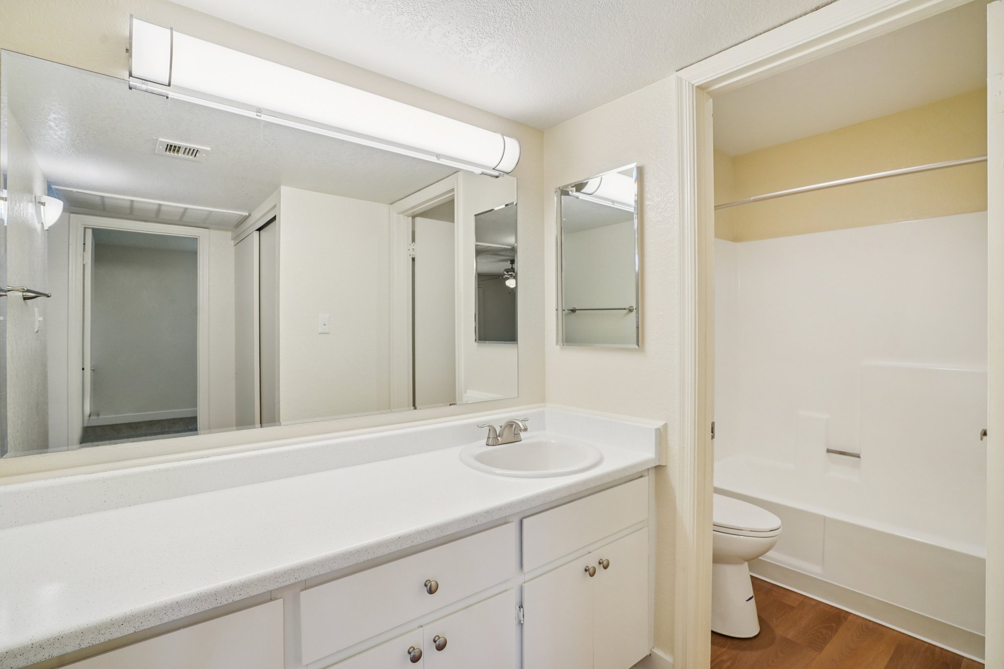 A clean, modern bathroom featuring a white countertop with a sink, a large mirror above, and white cabinetry. On the right, there is a toilet and a shower area with a simple white tub. The walls are light-colored, and the flooring is a warm wood finish. Good natural lighting is present.