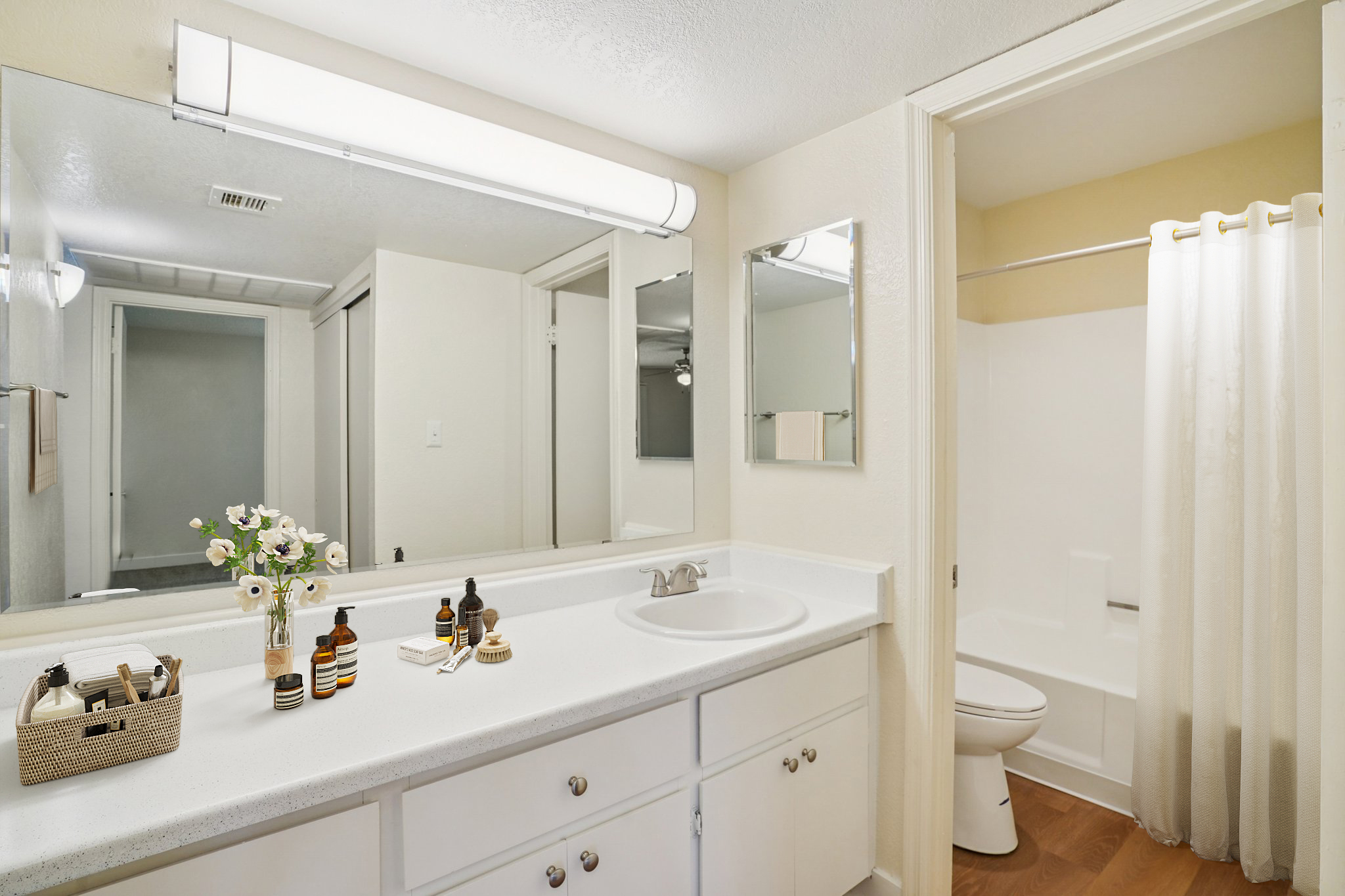 Bright and modern bathroom featuring a large mirror, white countertop, and a clean bathtub with a shower curtain. Decorative items include a small basket and various skincare products arranged neatly on the counter. The overall design is simple and inviting, with neutral colors and good lighting.