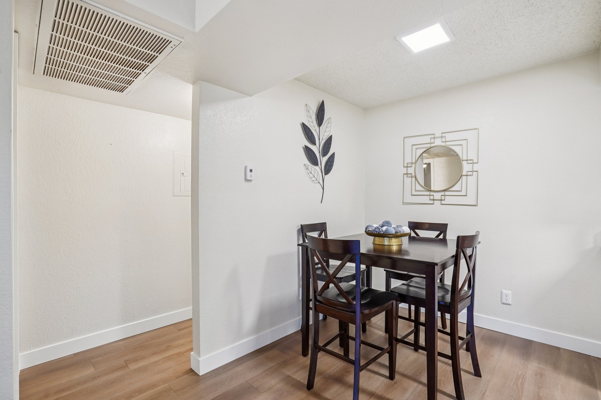 A small dining area featuring a rectangular table with four dark wooden chairs. The table has a decorative bowl in the center. The walls are painted white, with a minimalist wall art piece featuring a mirror and a leaf design. A doorway leads to another room, and there is a ceiling vent visible.