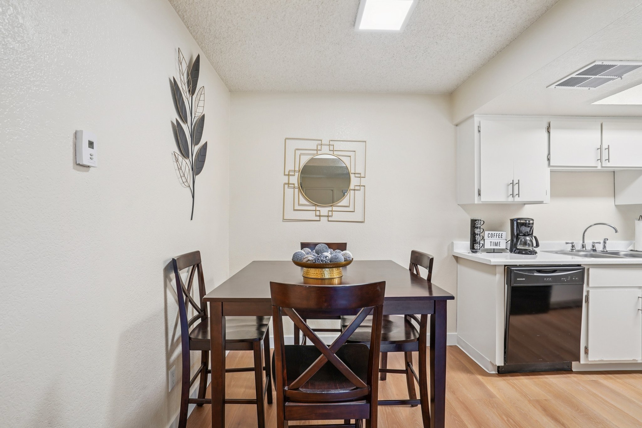 A modern kitchen dining area featuring a small wooden table with four tall chairs, a decorative bowl centerpiece, a round mirror on the wall, and a potted plant. The kitchen is equipped with white cabinets and appliances, and has a light-colored wall. Natural light enters through a ceiling fixture.